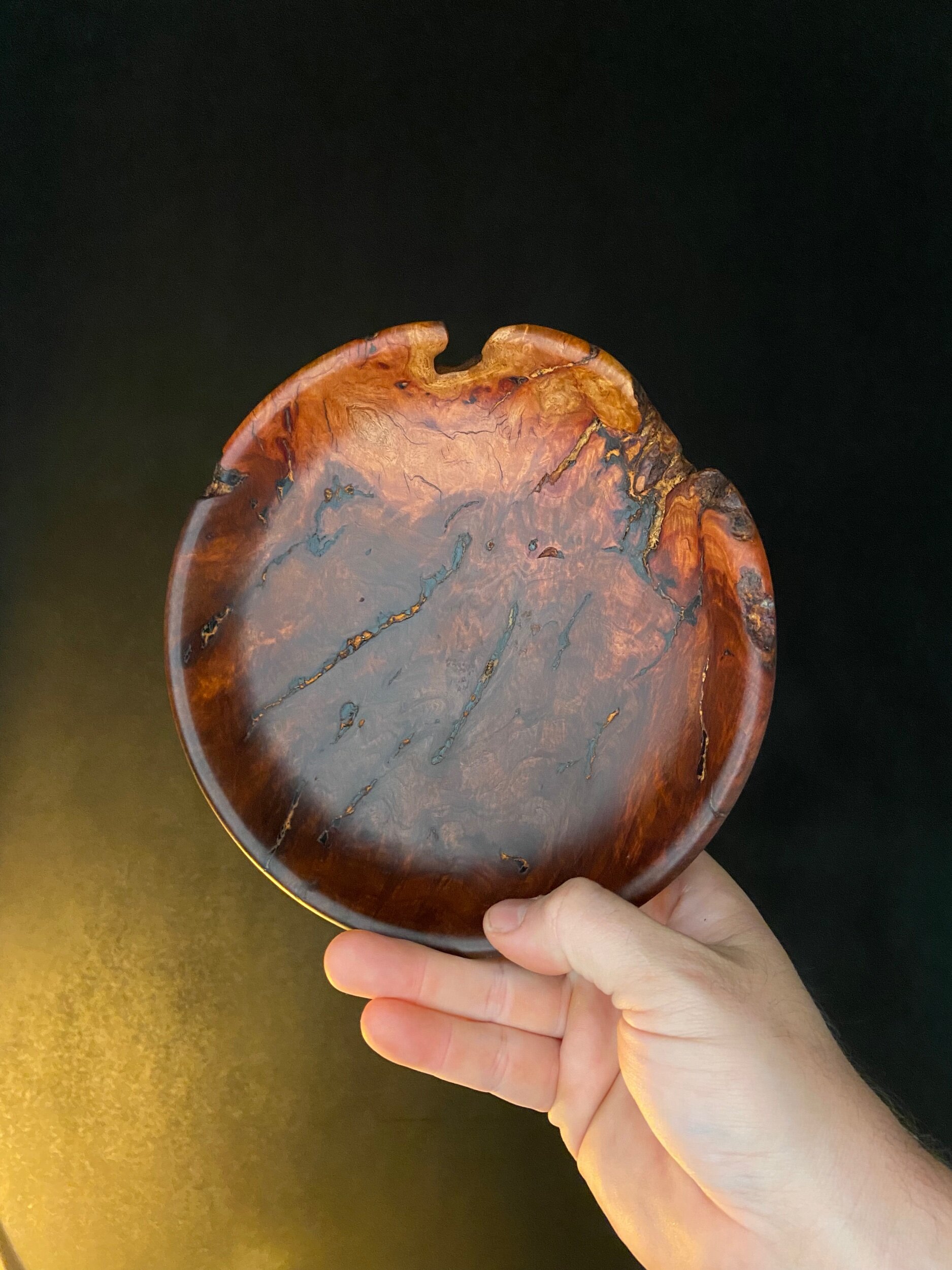 Hand holding a round wooden bowl with a glossy finish and natural wood grain patterns against a black background.