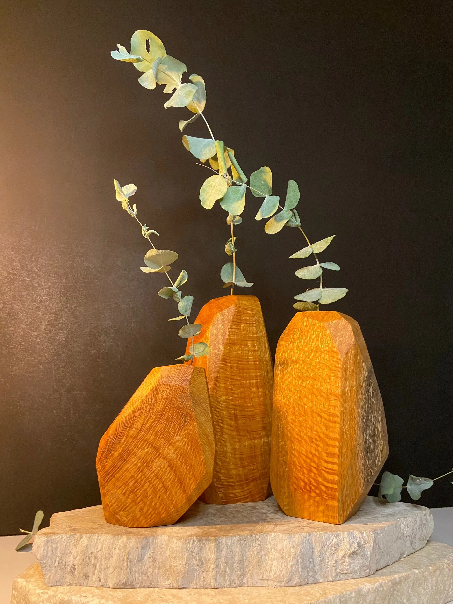 Decorative wooden vases with eucalyptus branches on a stone slab against a dark background.