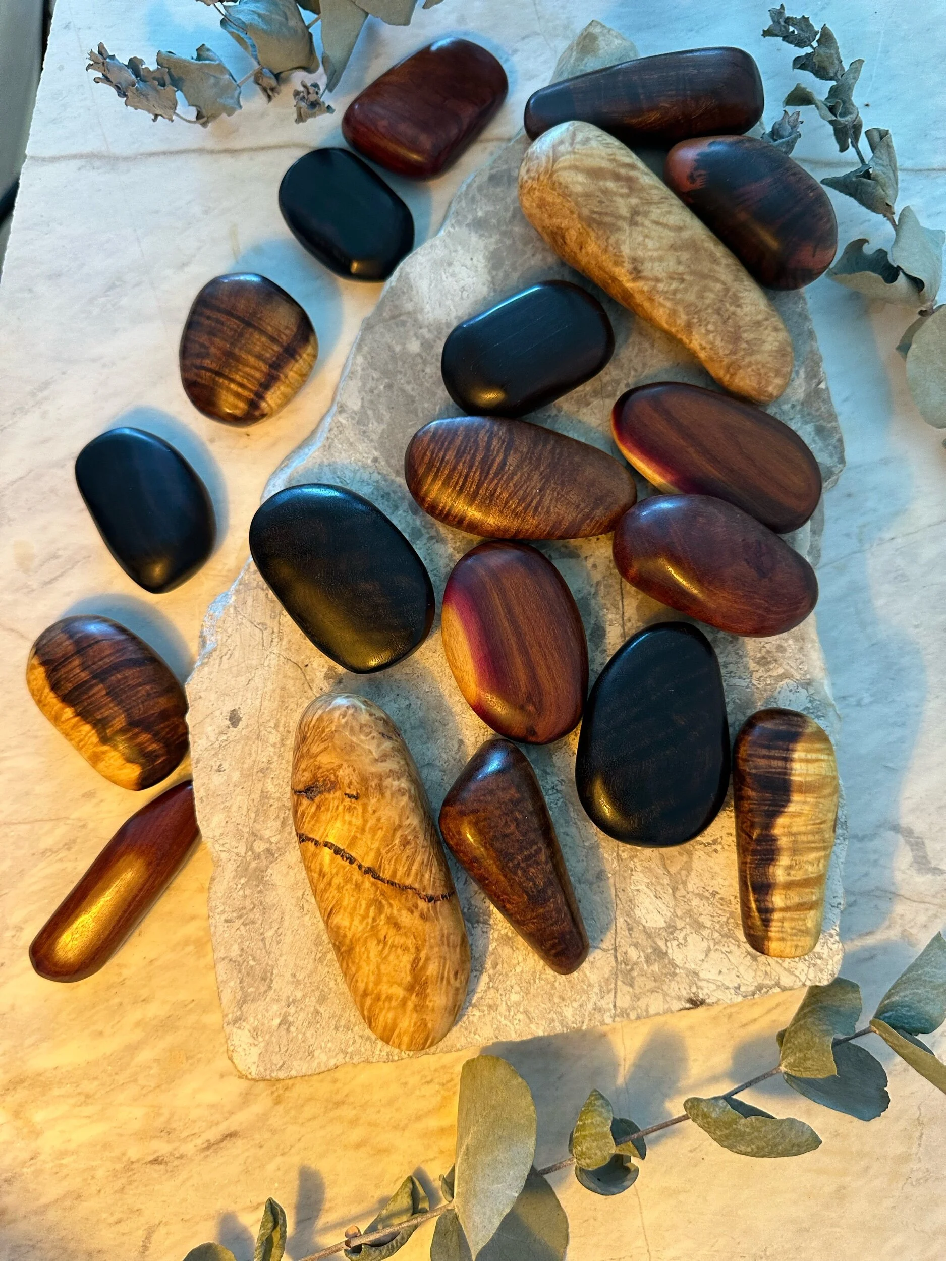 Various polished wooden stones of different shapes and sizes arranged on a light-colored stone surface with dried eucalyptus leaves around the edges.