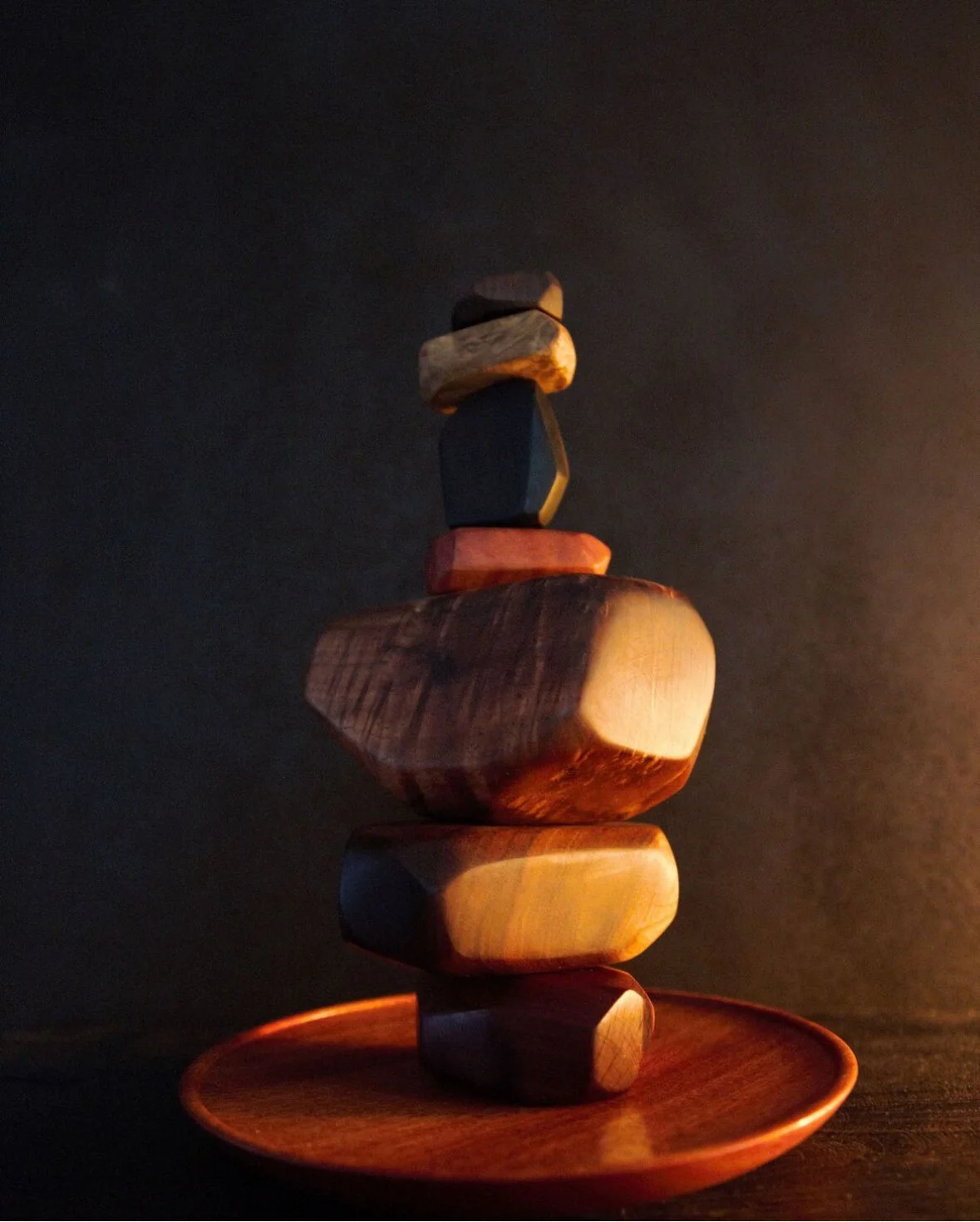 Stacked wooden stones of various shapes and sizes on a wooden plate, illuminated by warm lighting against a dark background.