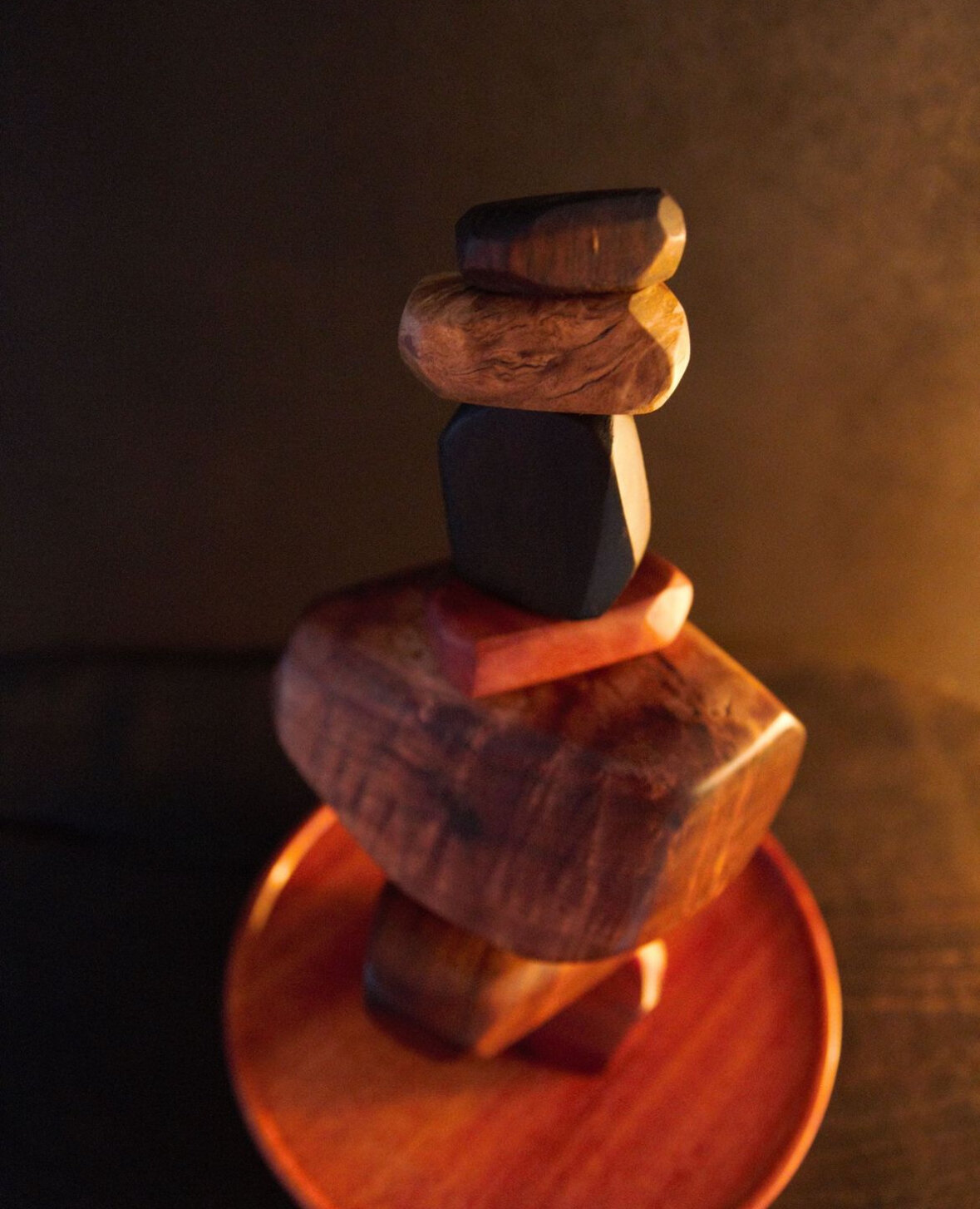 A stack of five irregularly shaped wooden blocks balanced on top of each other on a small round wooden platform with warm lighting.