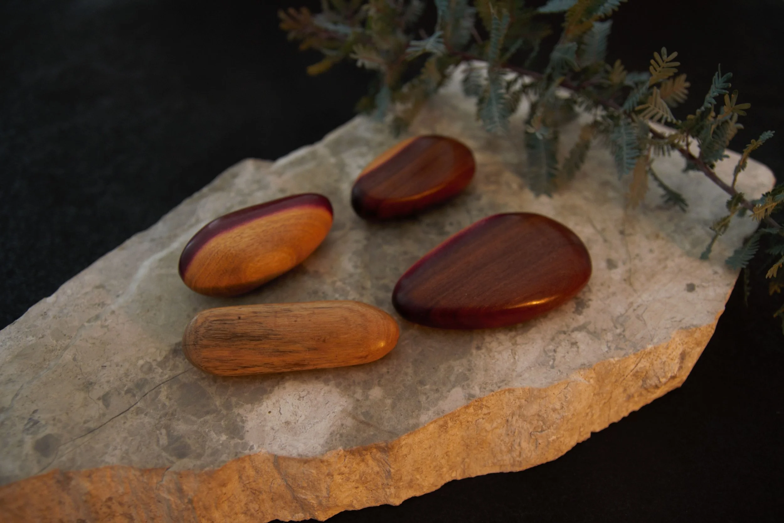 Four polished wooden stones of varying shapes and shades of brown placed on a textured light-colored stone slab with a sprig of greenery nearby.