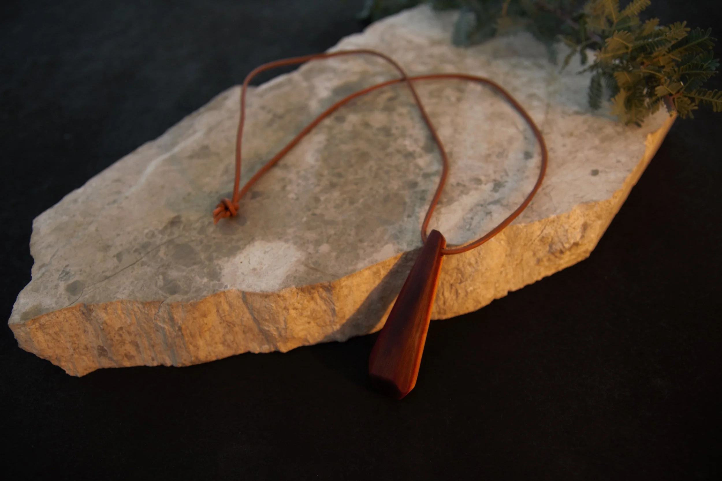 A wooden pendant necklace with a teardrop-shaped pendant on a leather cord, placed on a textured stone slab with some greenery.