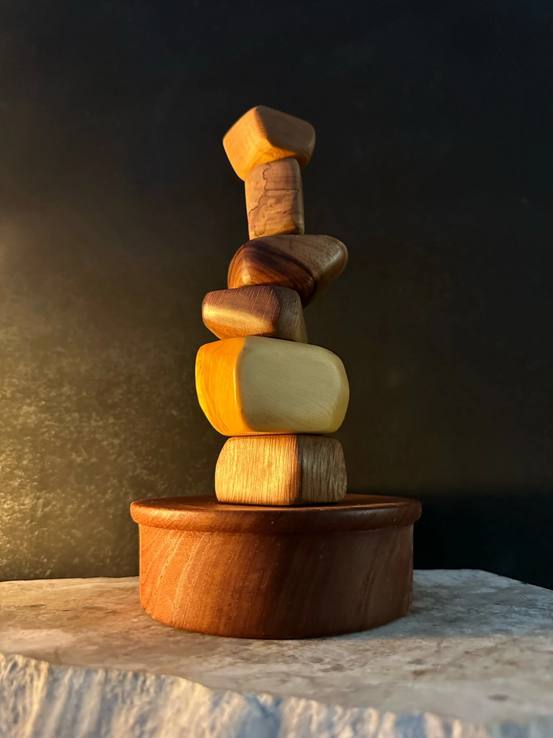 Stack of seven wooden blocks in various shapes and colors on a round wooden base, placed on a textured stone surface with a dark background.