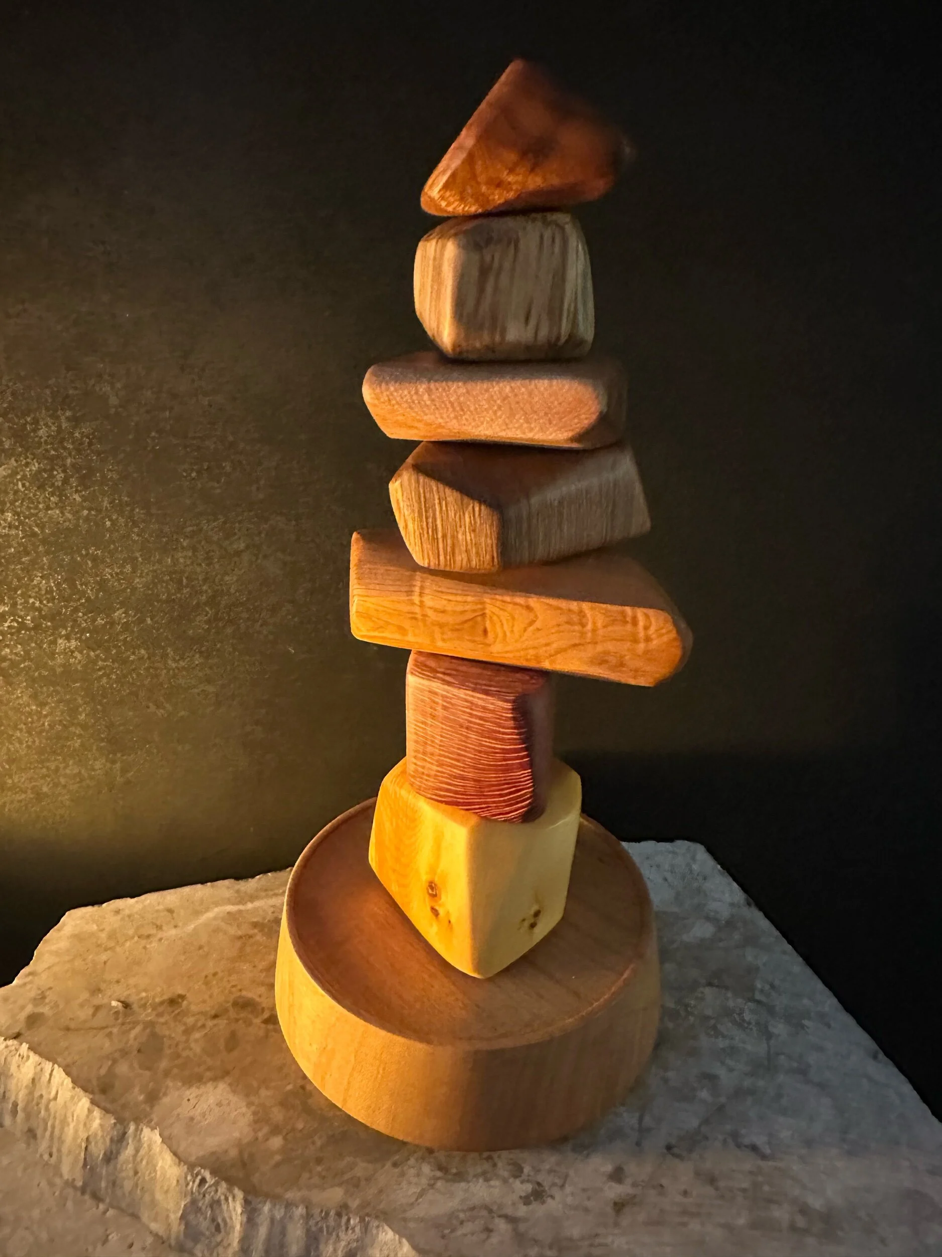 Stack of variously shaped wooden blocks balanced vertically on a round wooden base against a dark background.