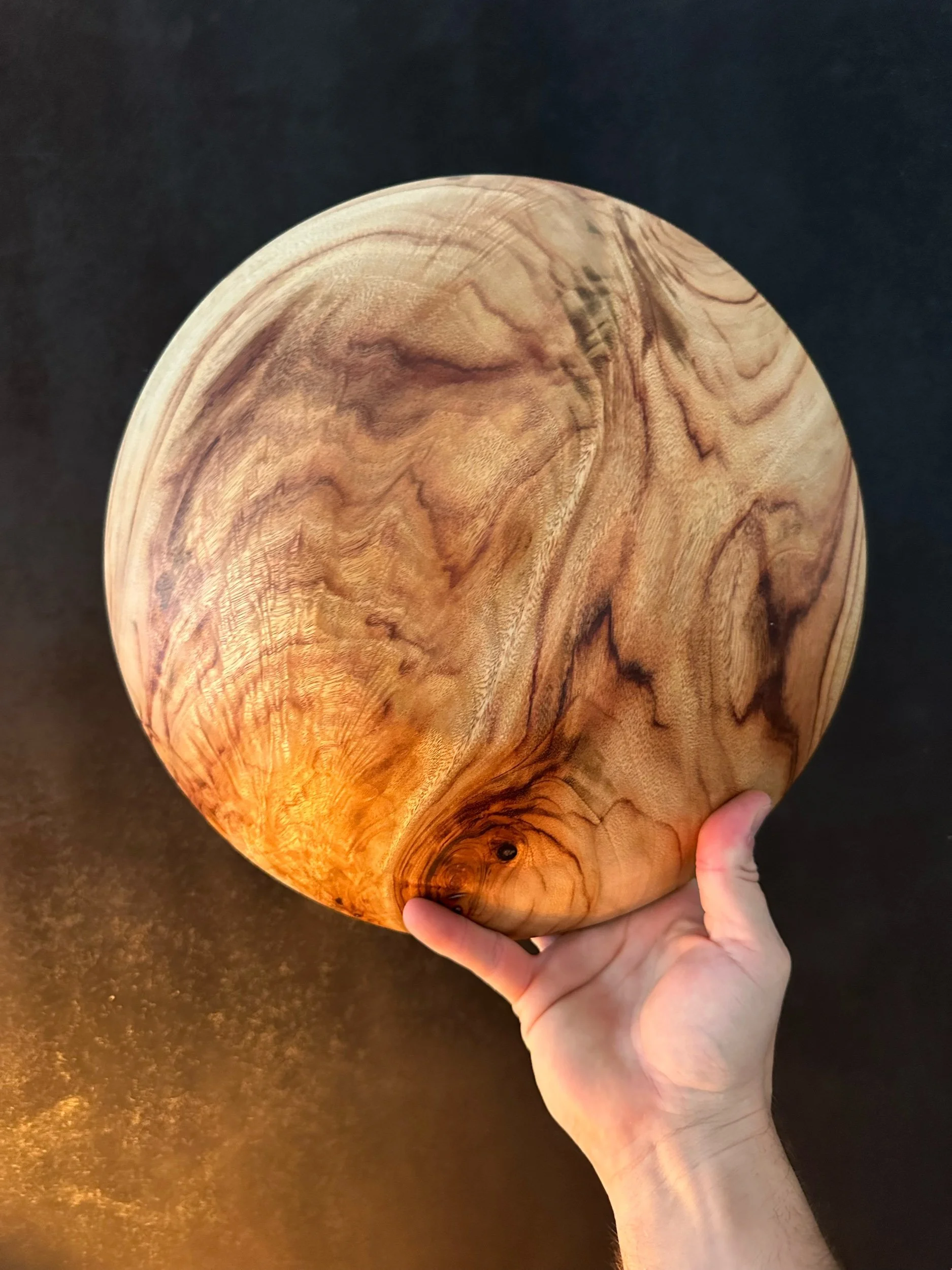 Hand holding a round wooden bowl with a natural wood grain pattern, against a dark background.