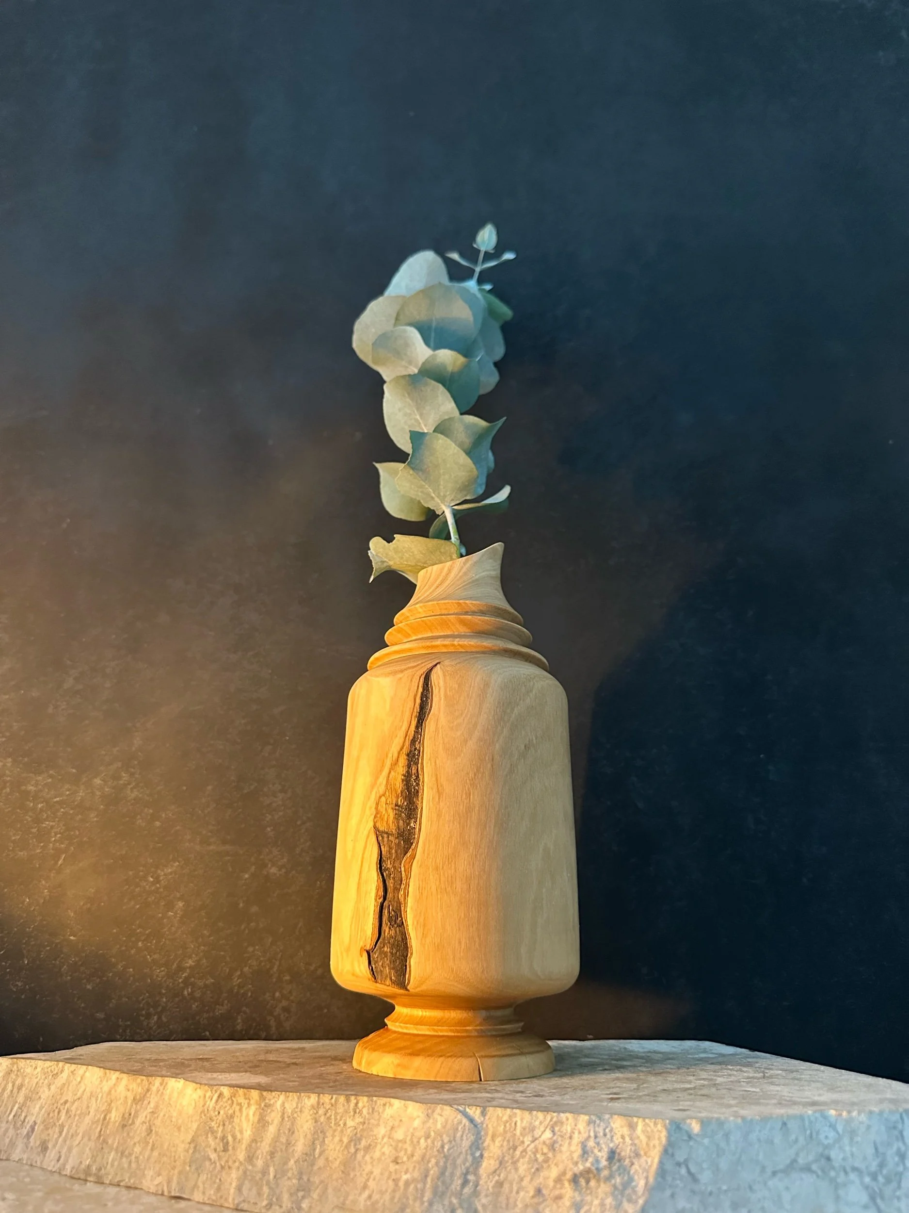 A modern wooden vase with a natural grain and black streak, holding a single sprig of eucalyptus, placed on a beige stone surface against a dark background.