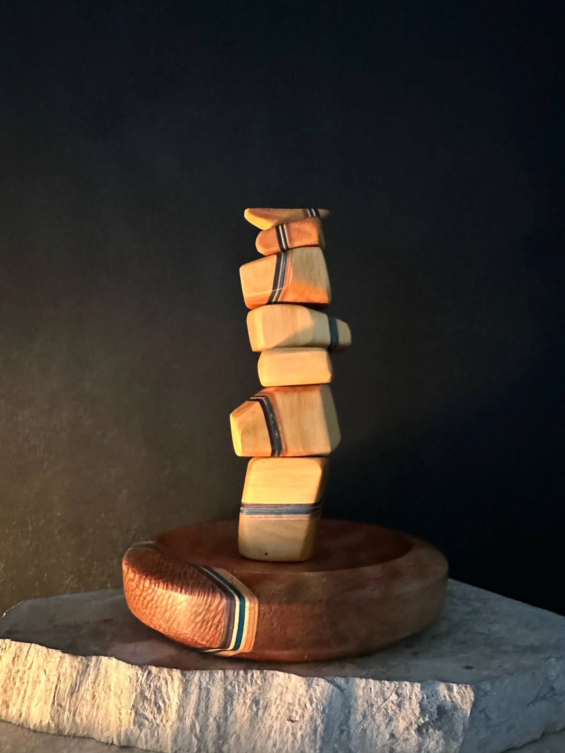 A vertical stack of wooden dominoes with multi-colored stripes balances on a round wooden base against a dark background.