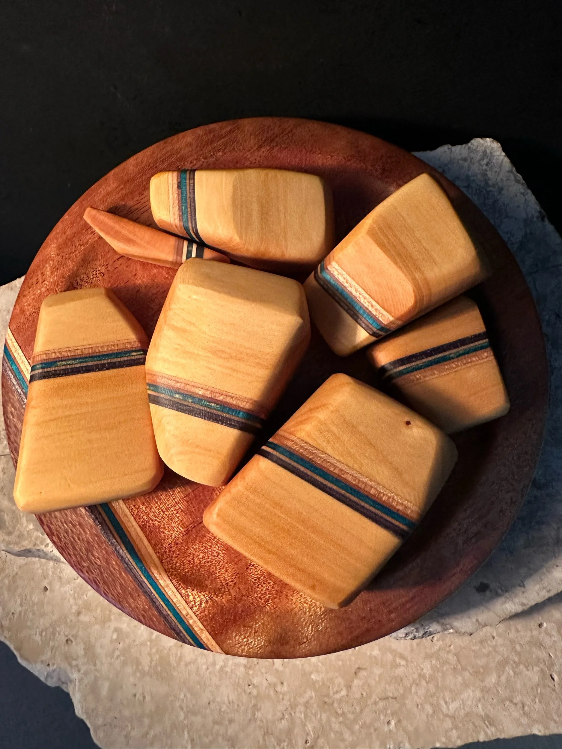 A round wooden bowl with nine rectangular blocks of wood inside, each with multicolored stripes near one end, on a textured surface against a dark background.