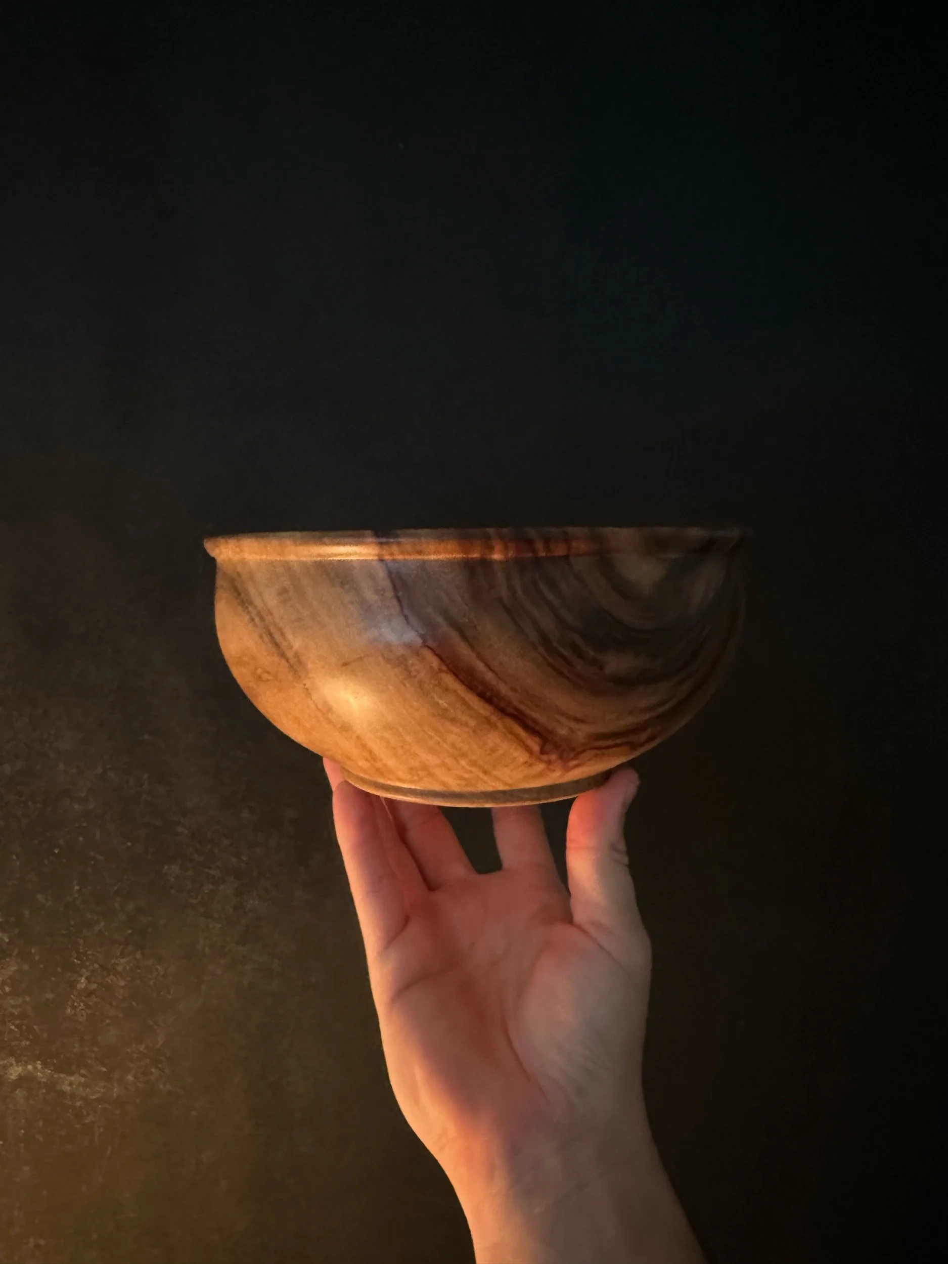 A hand holding a wooden bowl with a dark, swirling grain pattern against a dark background.