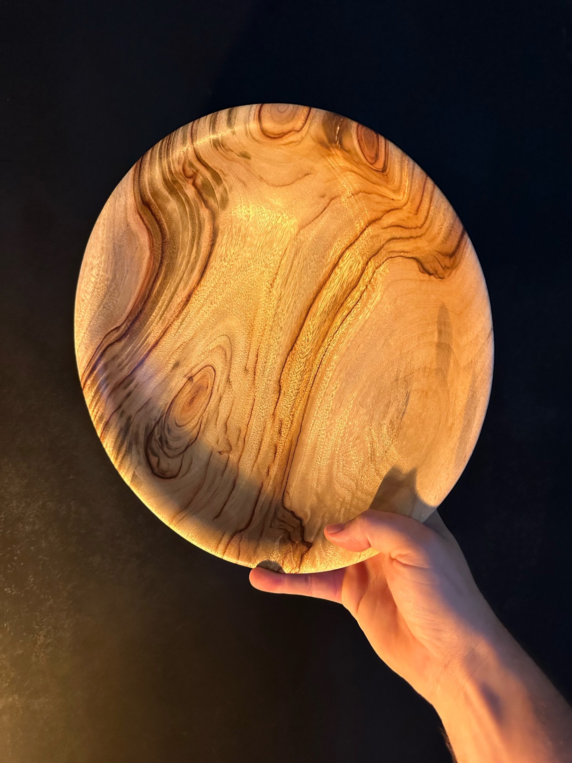 A hand holding a round wooden plate with intricate wood grain patterns against a dark background.