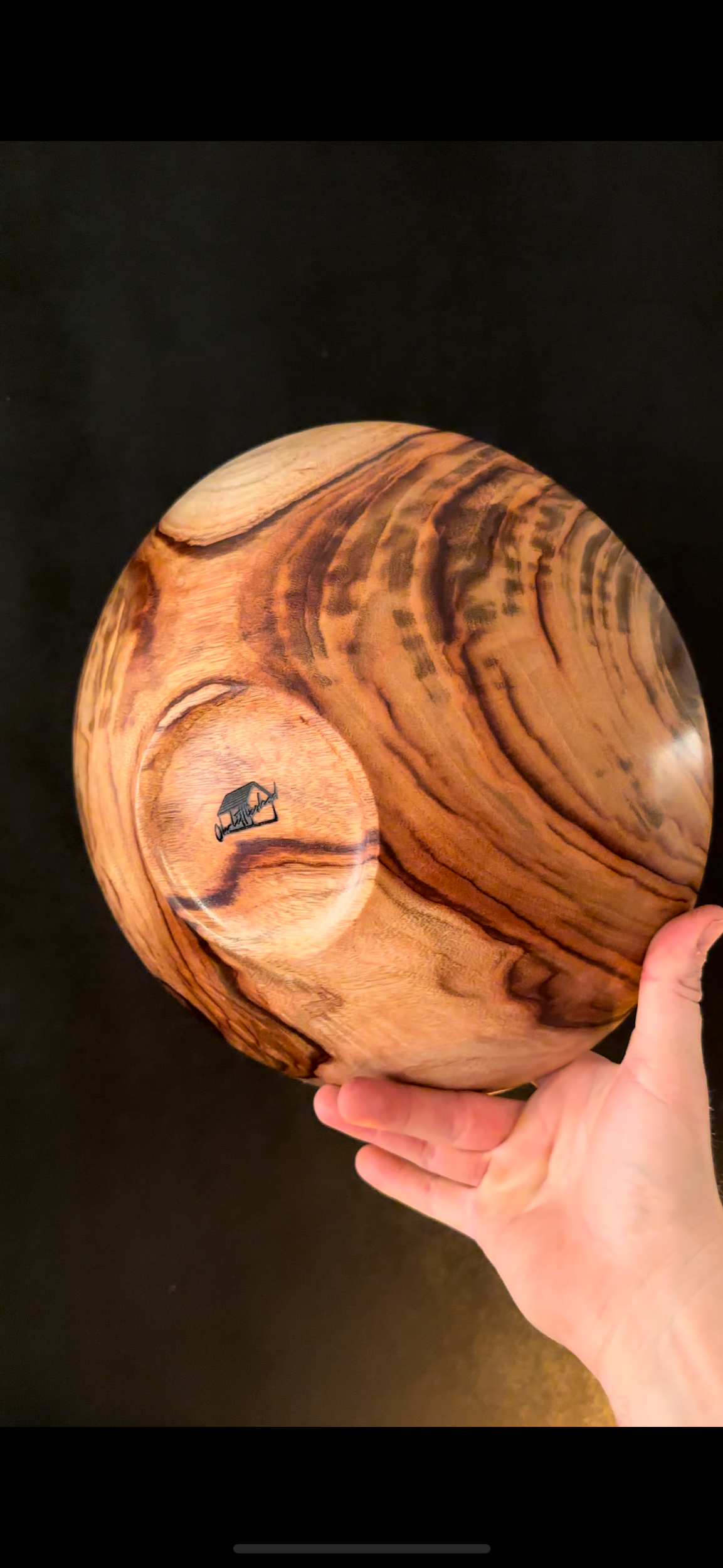 A hand holding a round wooden bowl with rich, swirling wood grain patterns and a small black logo inside the bowl.