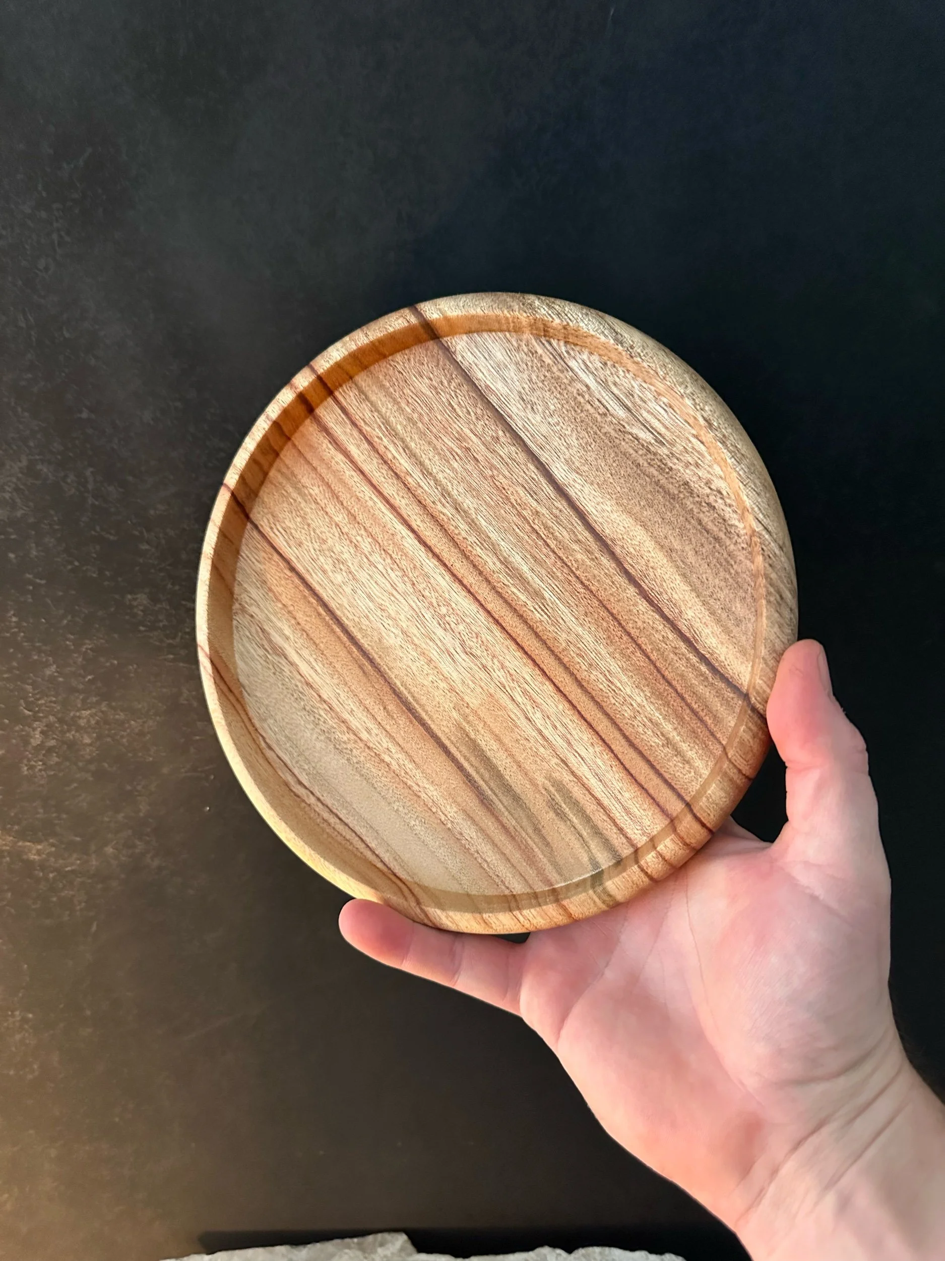 A round wooden plate with a natural wood grain pattern, being held in a person's hand over a dark surface.