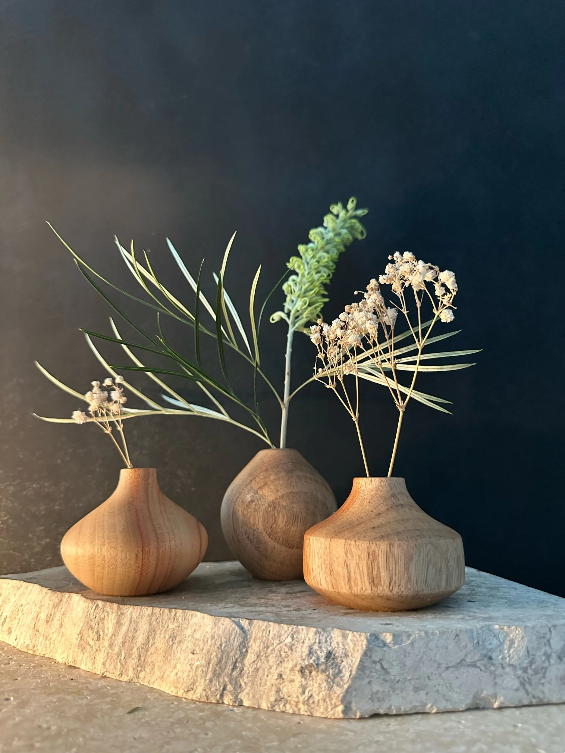 Three wooden vases with dried and green plants placed on a light-colored stone slab against a dark background.