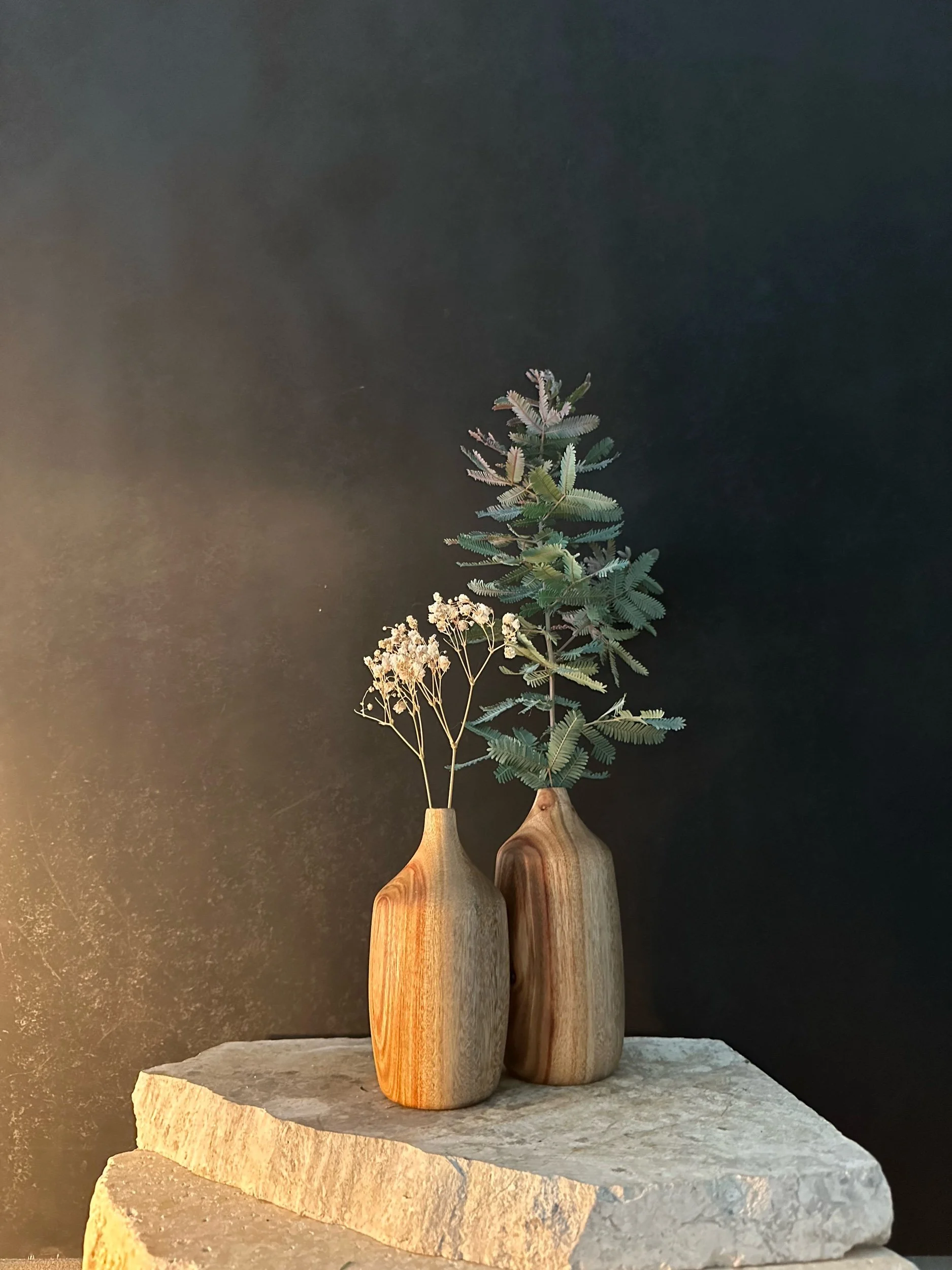 Two wooden vases with green foliage and dried flowers, sitting on a stone slab against a dark background.