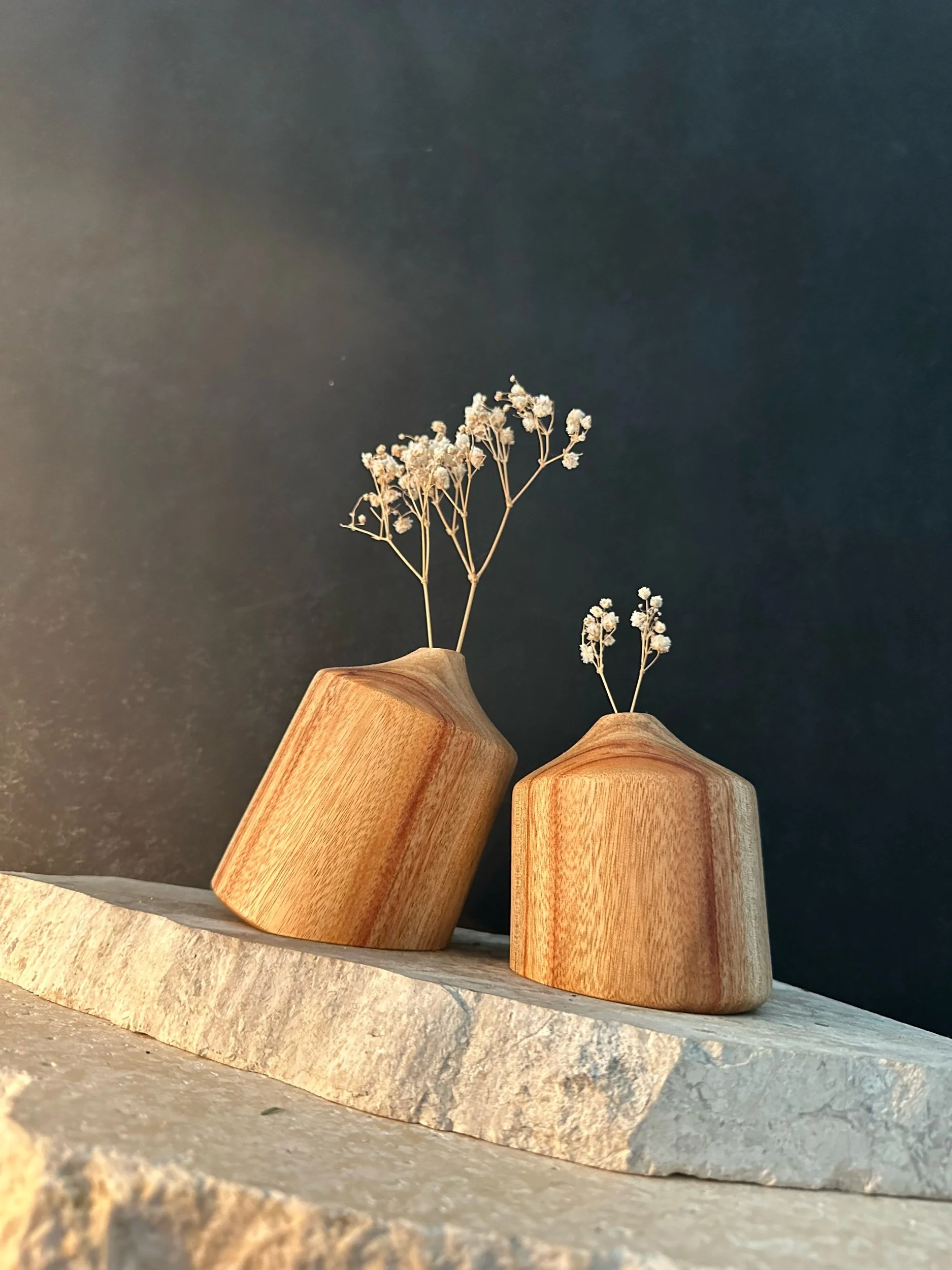 Two wooden vases with dried white flowers on a beige stone surface against a dark background.