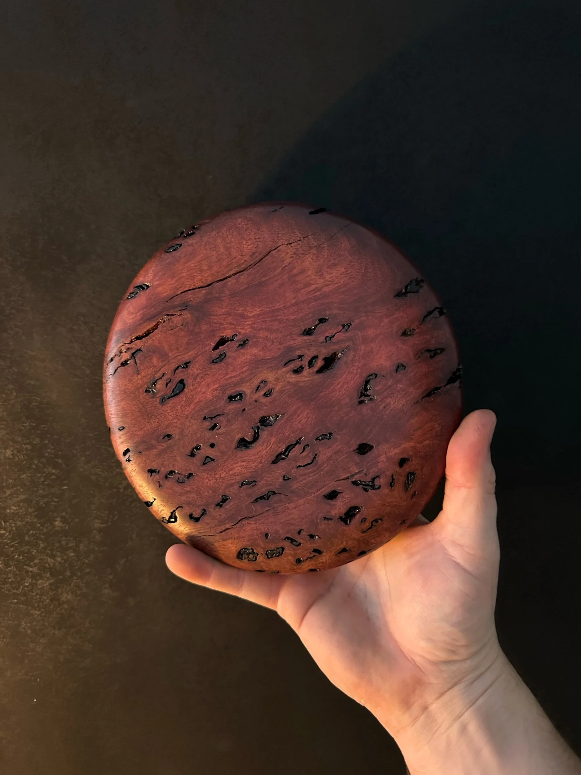 Hand holding a polished, round wooden object with a dark, grainy surface and natural cracks.
