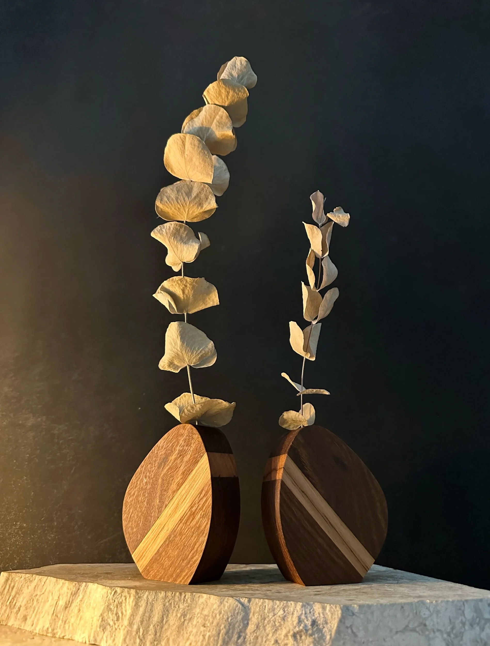 Decorative wooden vases with dried eucalyptus branches on a textured light-colored surface against a dark background.