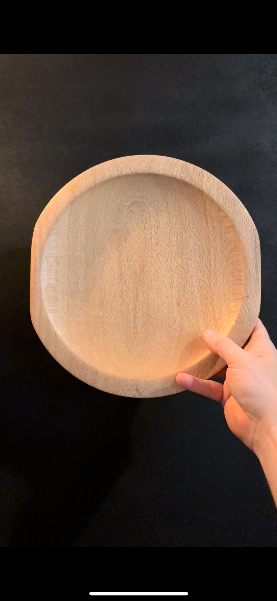 A person holding a round, unfinished wooden plate or tray against a black background.