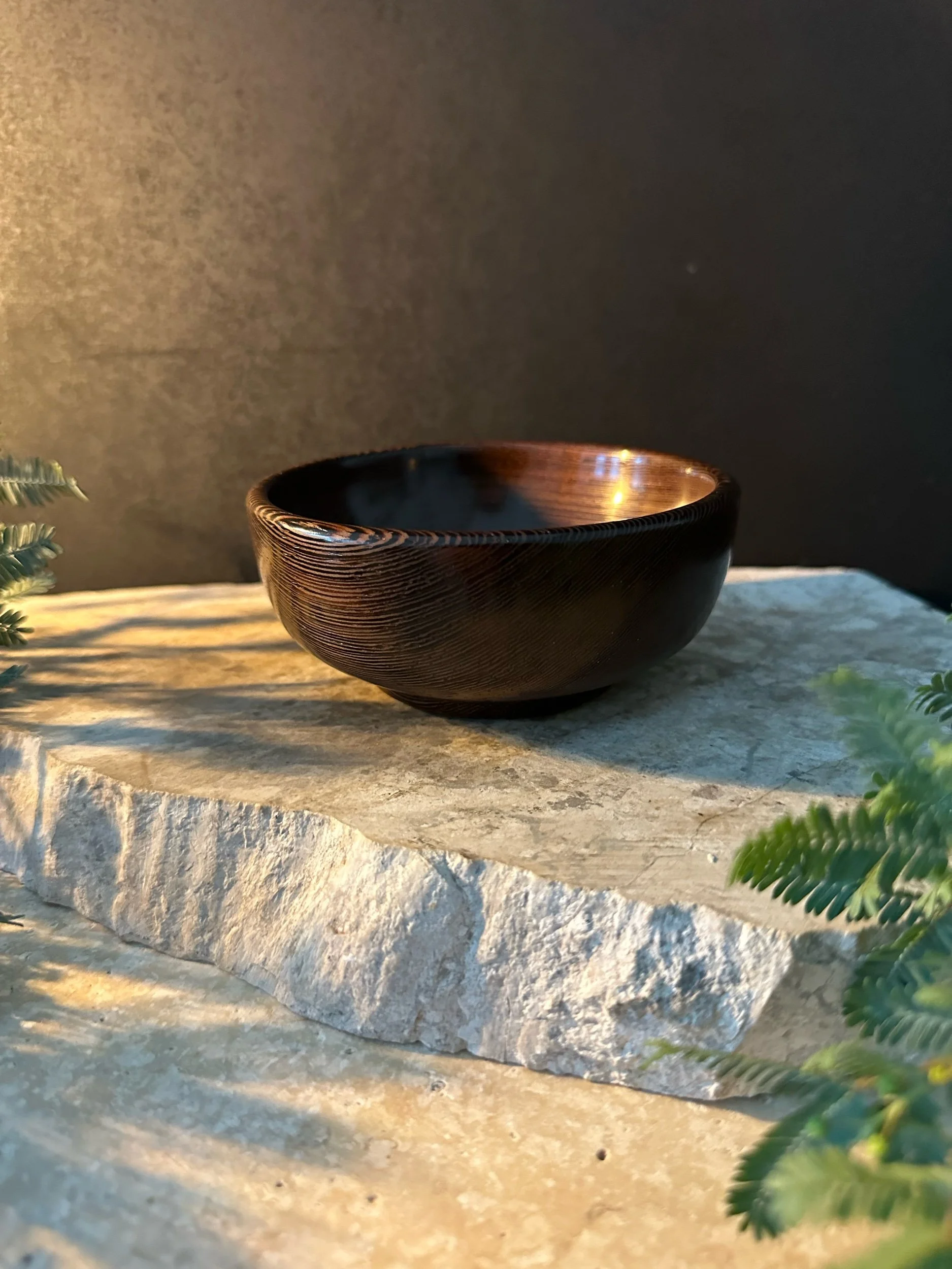 A dark wooden bowl with a shiny interior resting on a rough, light-colored stone surface, decorated with small green plants on the sides.