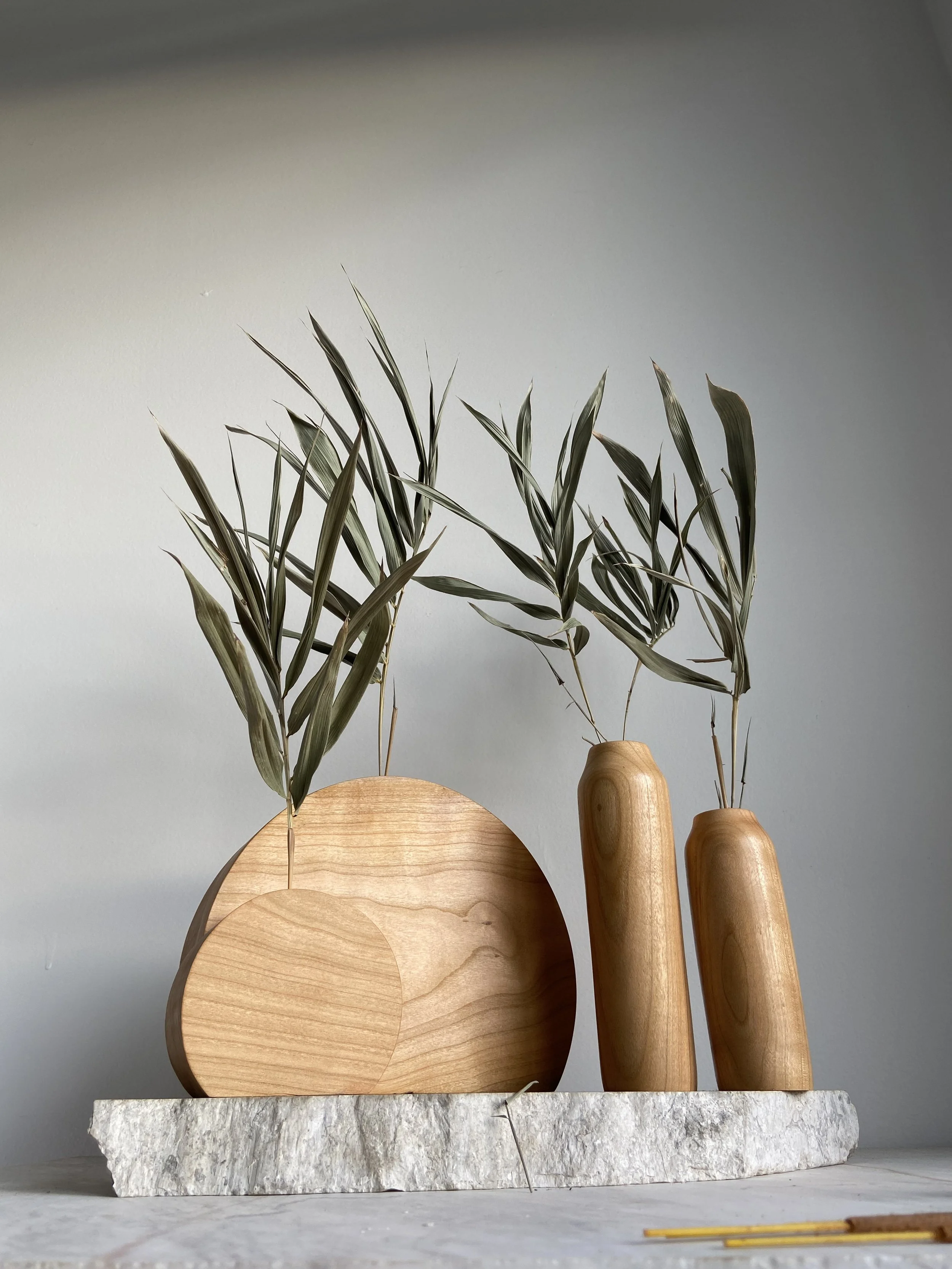 Decorative arrangement featuring two wooden vases with dried branches and leaves on a stone slab, against a plain light gray wall.