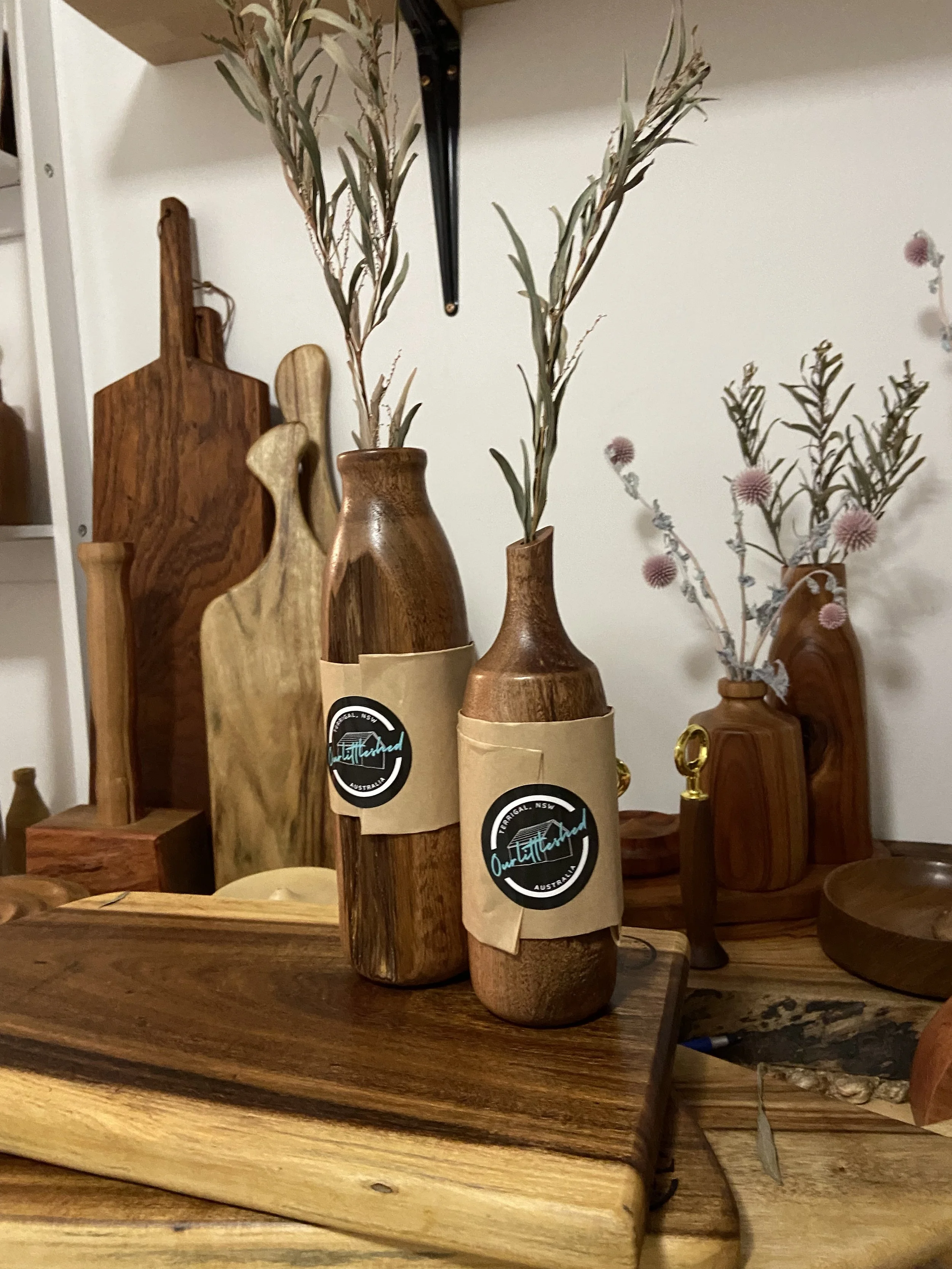 Two wooden vases with dried branches and leaves, each with a black and white label, on a wooden table with various wooden decorative items in the background.