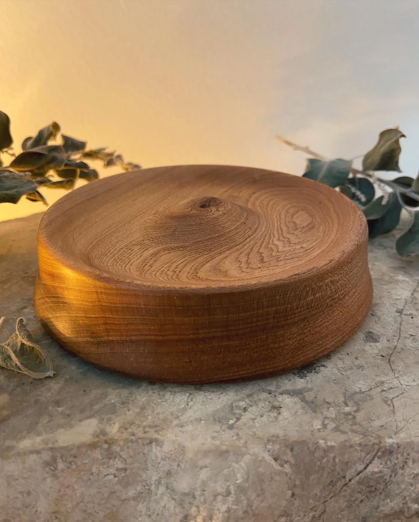 A round wooden platform with a smooth, polished surface and natural wood grain, placed on a beige marble surface with some dried leaves in the background.
