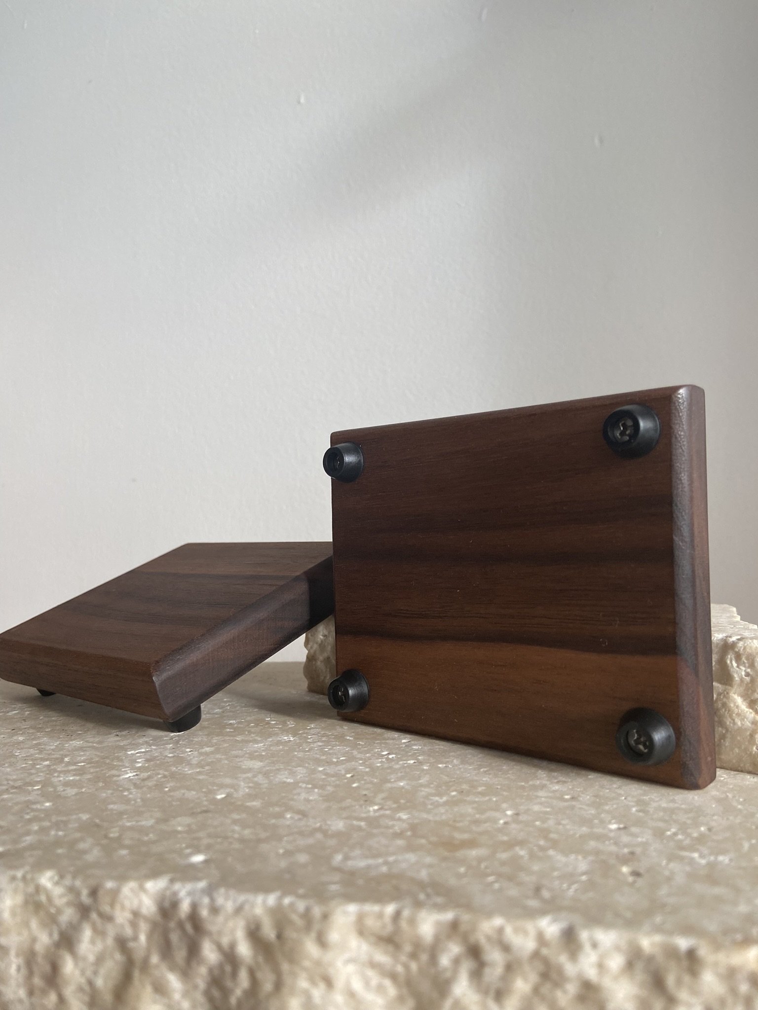 Close-up of a wooden object with a flat, square surface and rounded edges, resting on a rough, beige surface against a plain white wall. The object appears to be a piece of furniture or decorative item, with four visible screws on one side.
