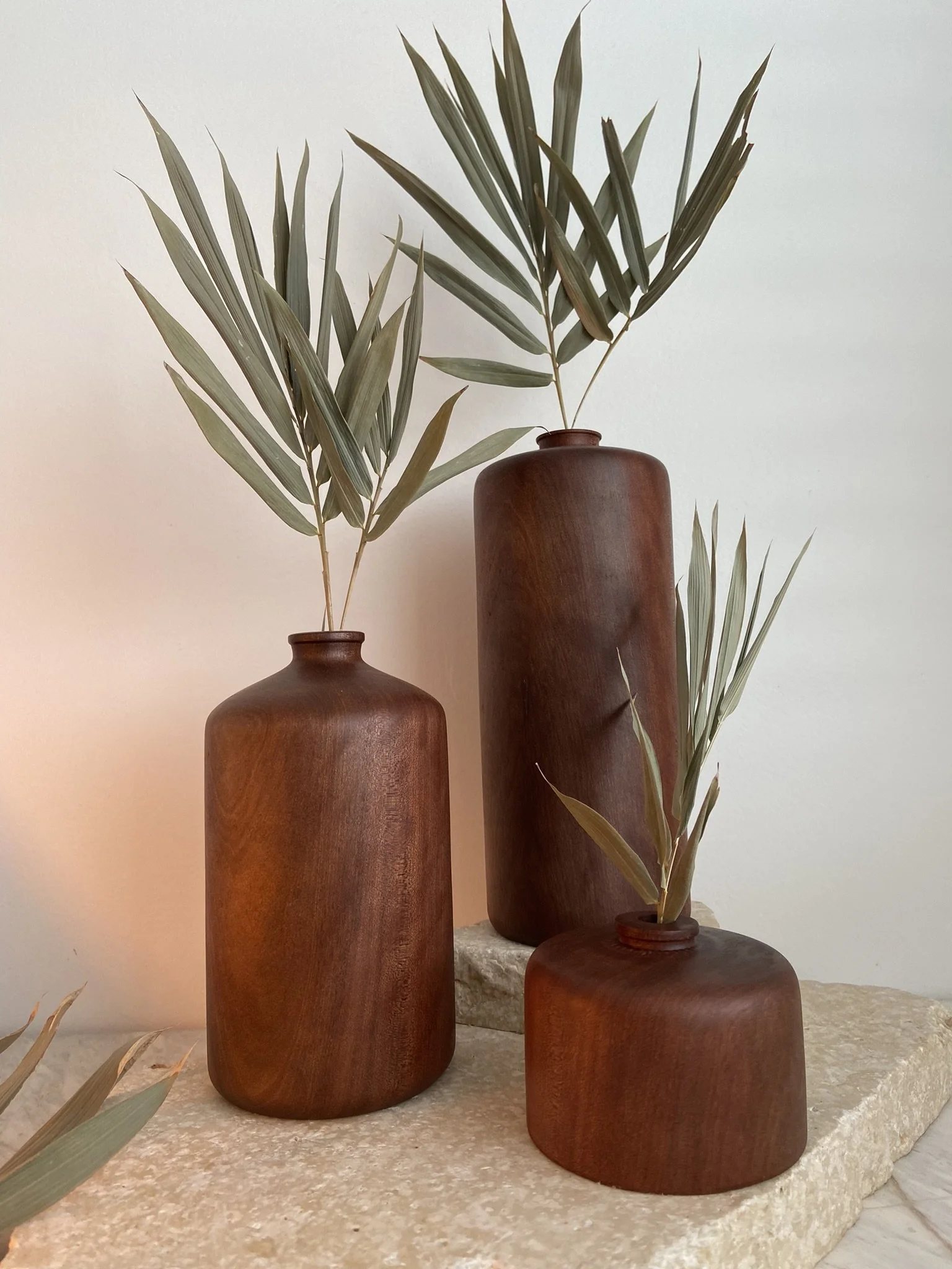 Three wooden vases with green leafy branches on a textured stone surface against a plain wall.