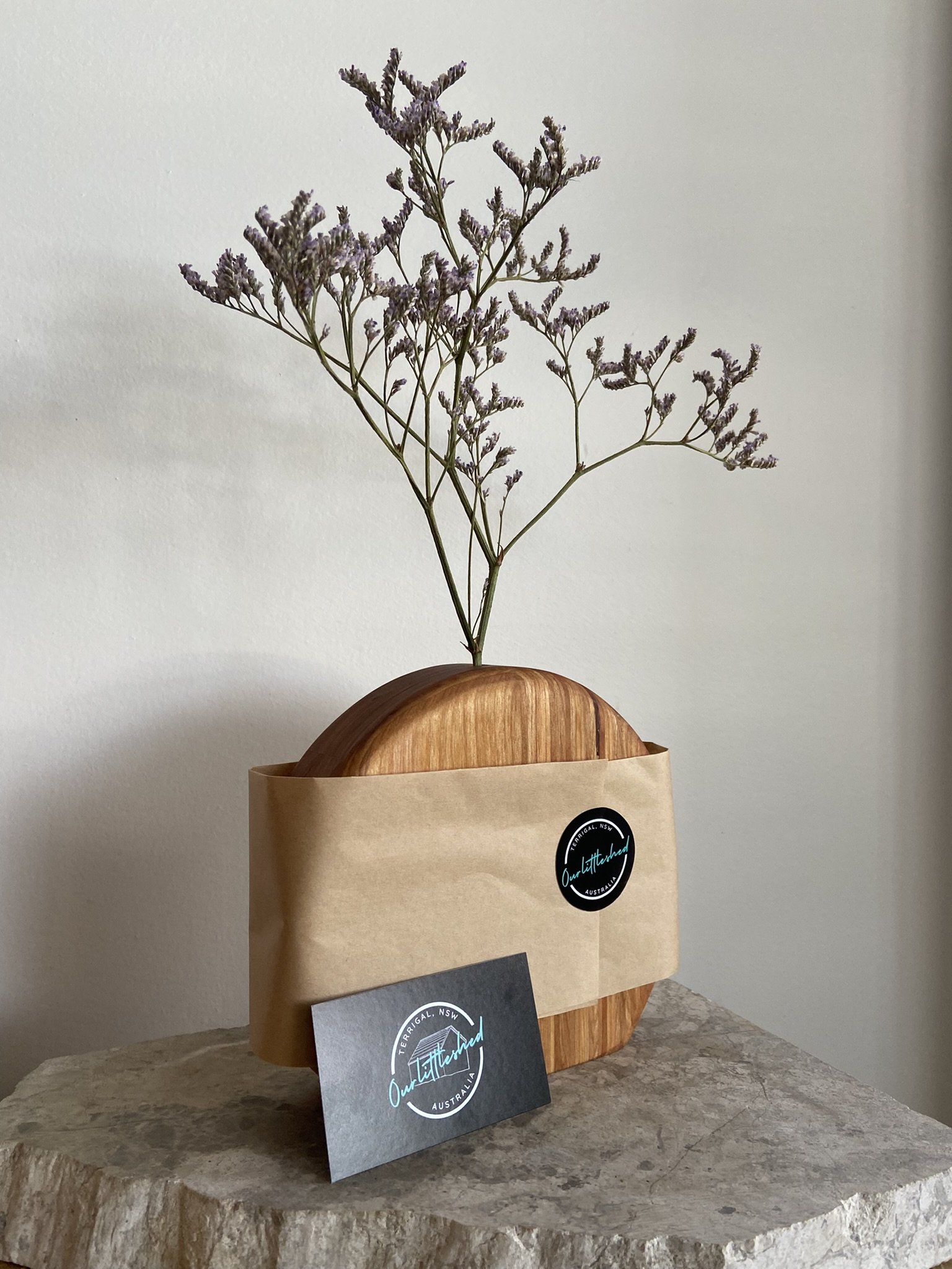 A minimalistic arrangement of a wooden object with a branch of small purple flowers, partially wrapped in a brown paper band, resting on a rough stone surface against a plain white wall.