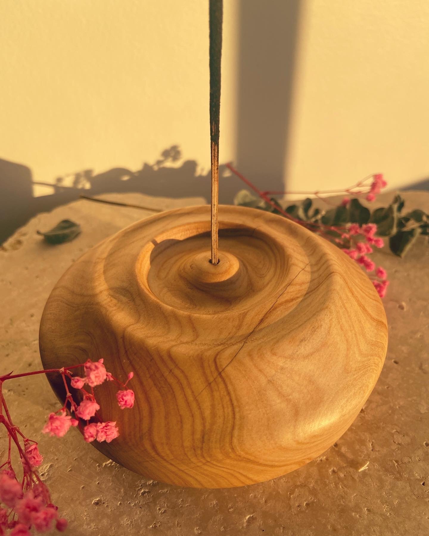 A round, wooden singing bowl with a wooden striker inside, placed on a textured surface with pink flowers in the background.