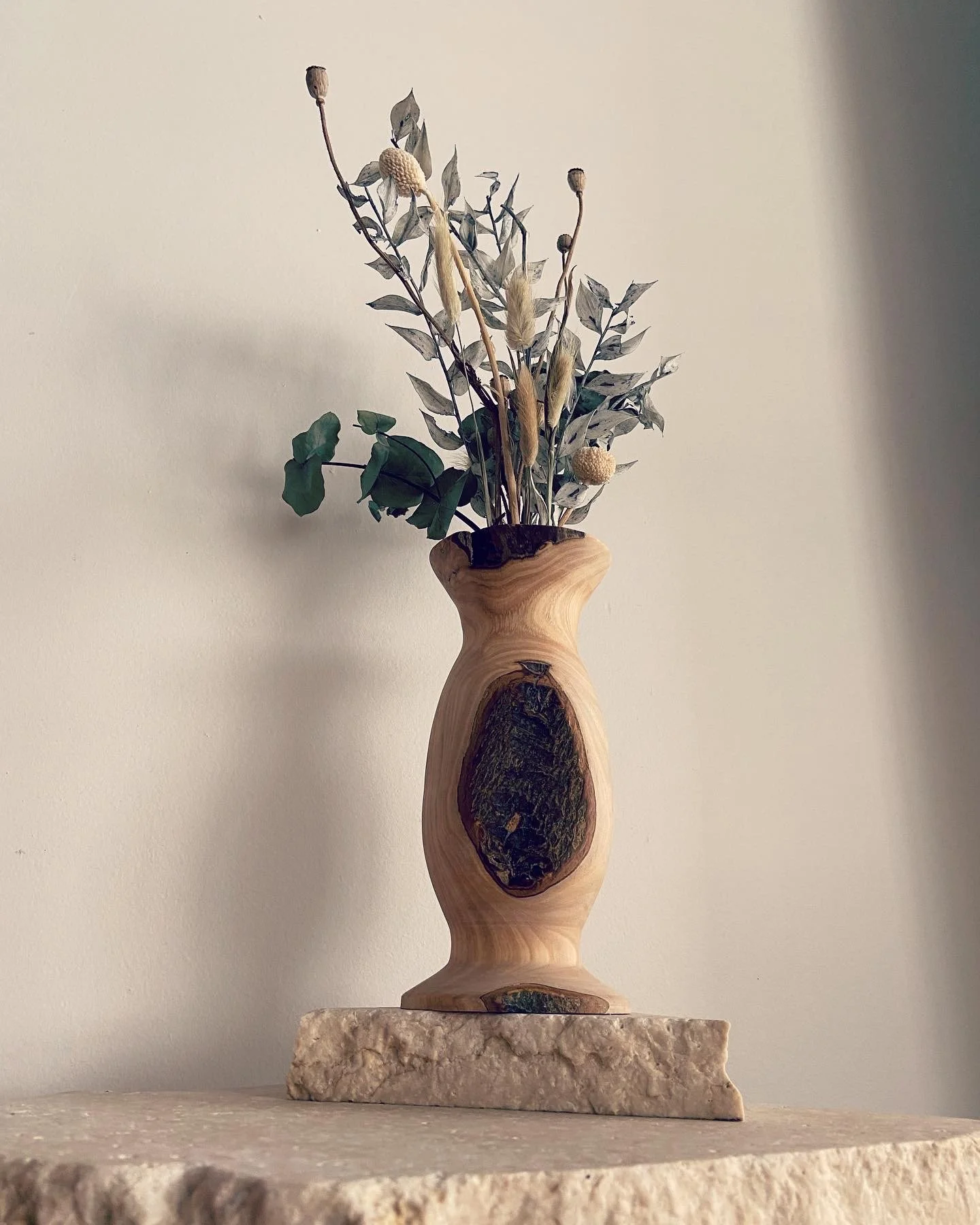 A wooden vase with a natural finish, holding dried flowers and greenery, placed on a stone surface against a plain wall.