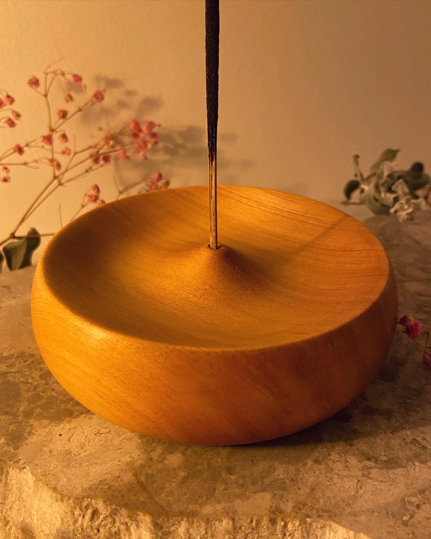 A wooden bowl with a metal rod in the center, set on a stone surface with flowers in the background.