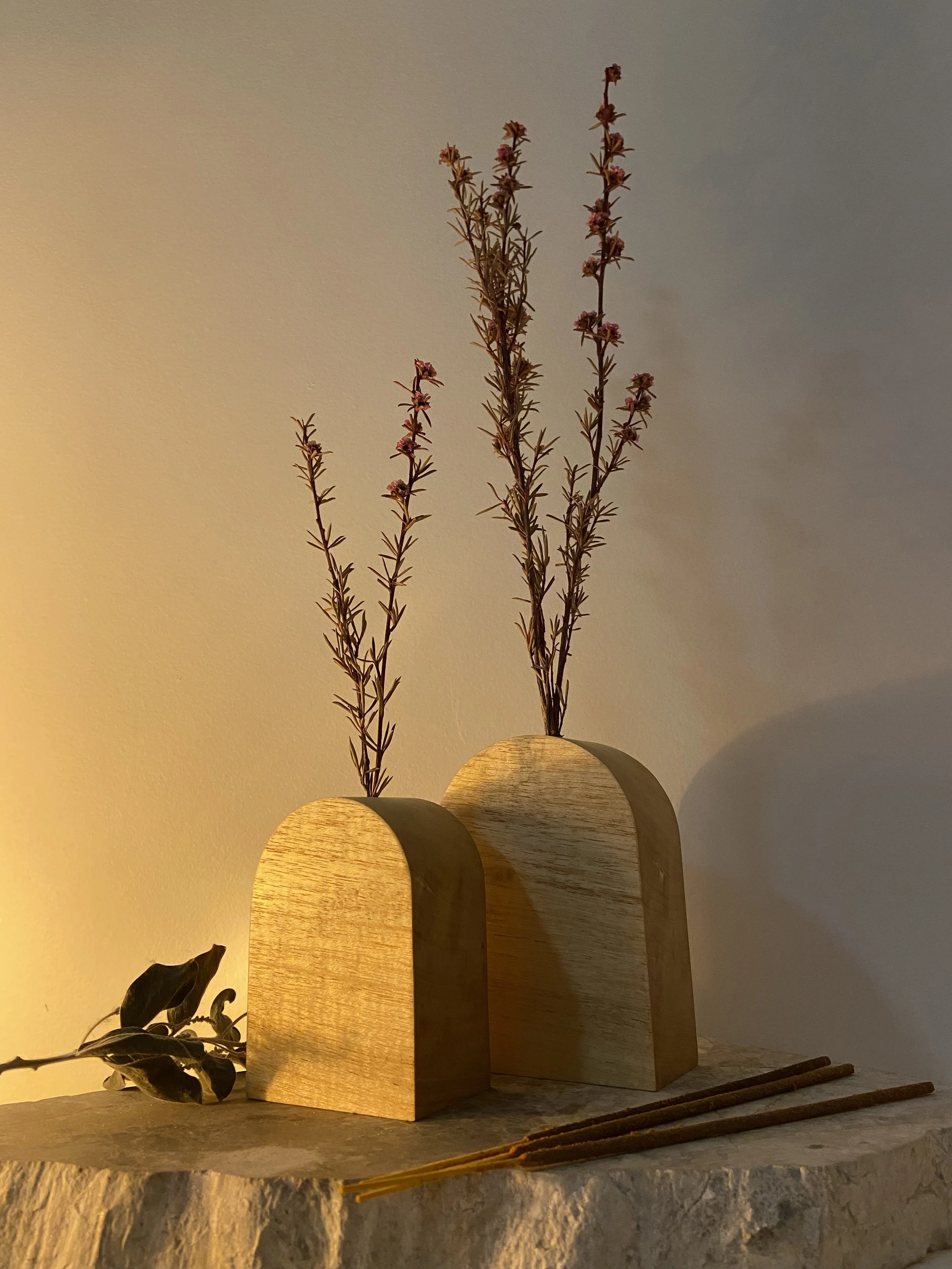 Decorative arrangement of two wooden vases with dried flowers and leaves on a stone surface, with a warm lighting.