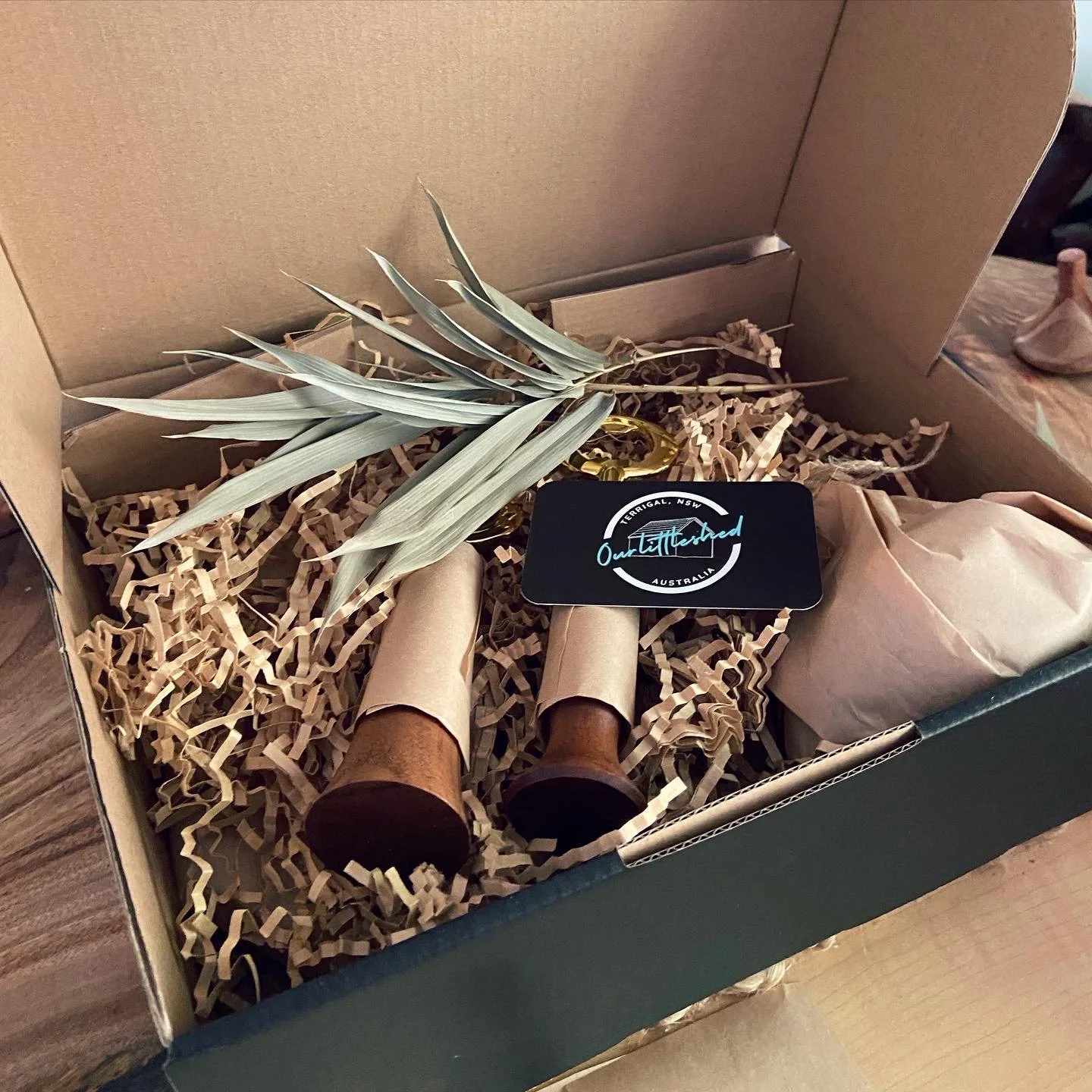 Open gift box containing a dried plant, two cardboard tubes, a black card, and a wrapped item, all nestled in shredded paper filler.