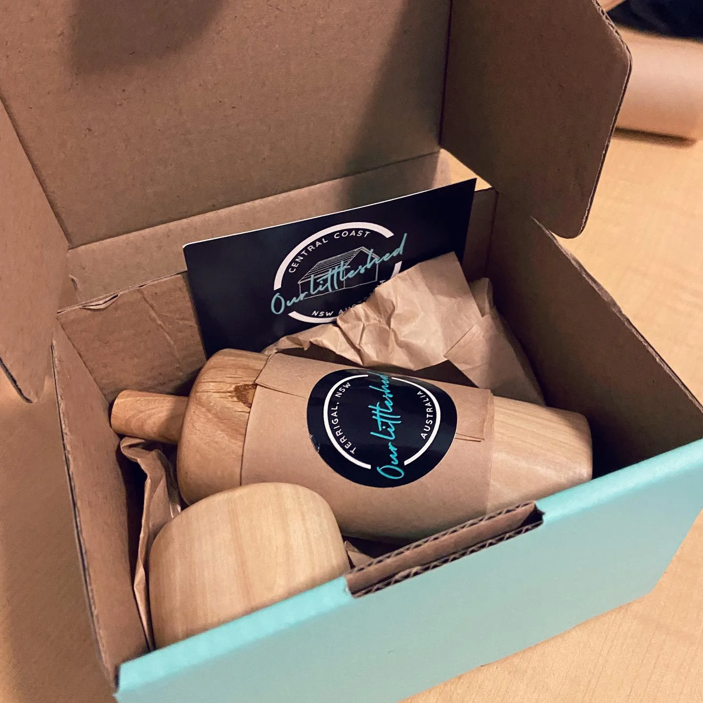 A cardboard box containing a wooden rolling pin with a sticker label from Out Littlered, a small container with a lid, and a business card from Out Littlered, all secured with crumpled brown paper inside the box.