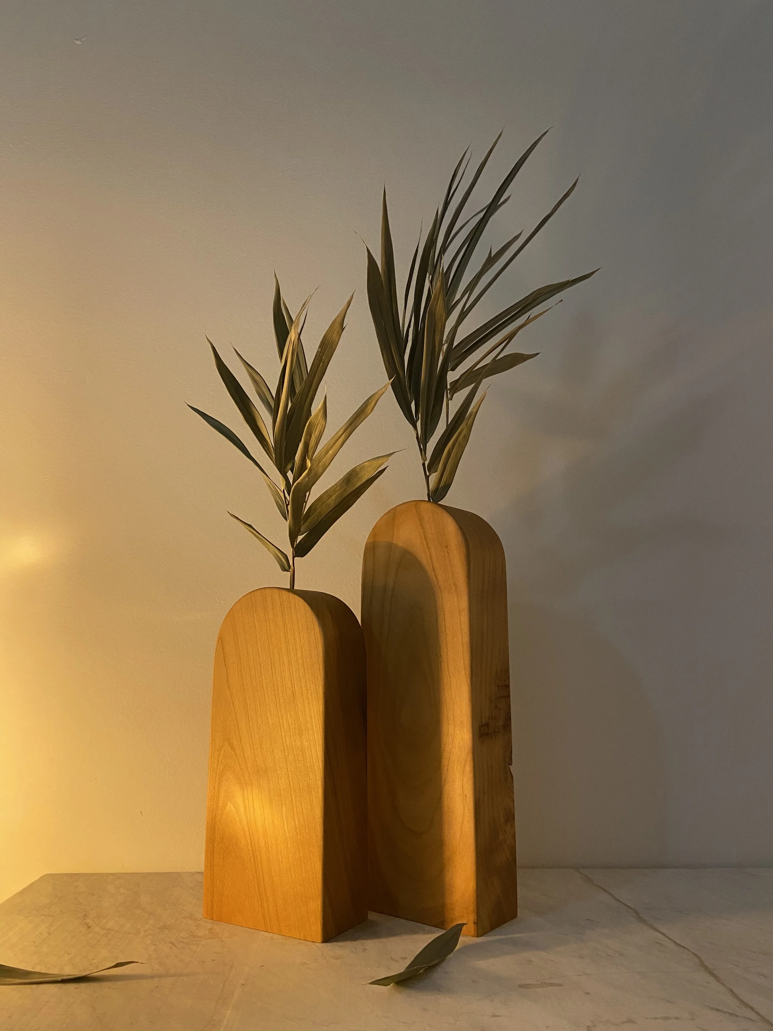 Decorative wooden vases with leafy branches on a light-colored surface against a neutral wall.
