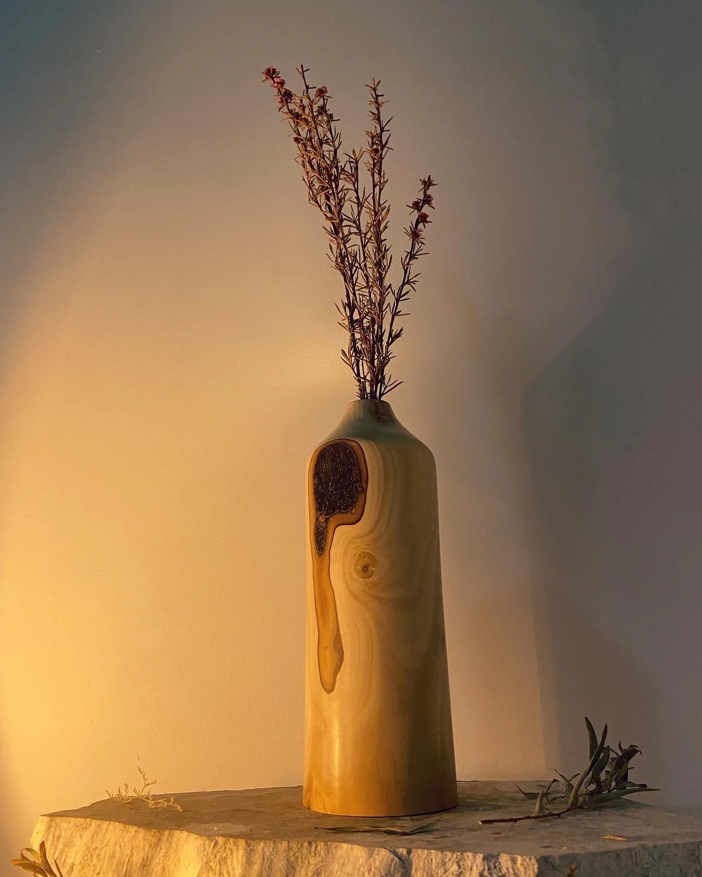 A wooden vase with a natural wood grain pattern, holding dried branches with small leaves, placed on a textured surface against a warm, softly lit background.