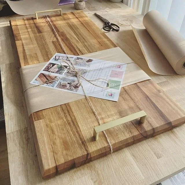 A wooden work table with a large cutting board, scissors, a roll of paper, and a package wrapped with kraft paper, string, and greeting cards.