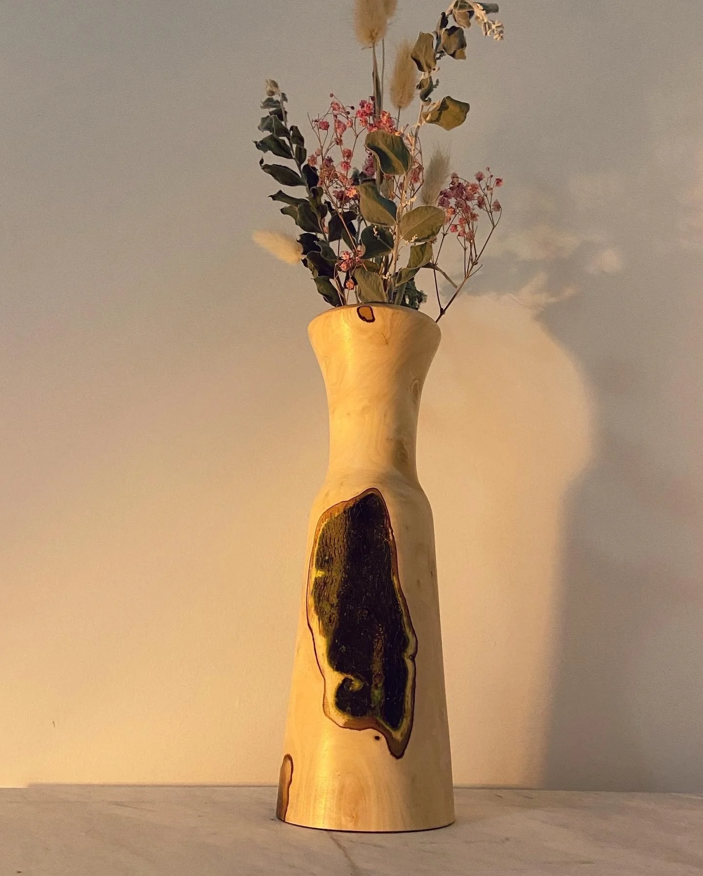 Wooden vase with natural knots and patterns, filled with dried flowers and foliage, placed on a surface against a plain wall.