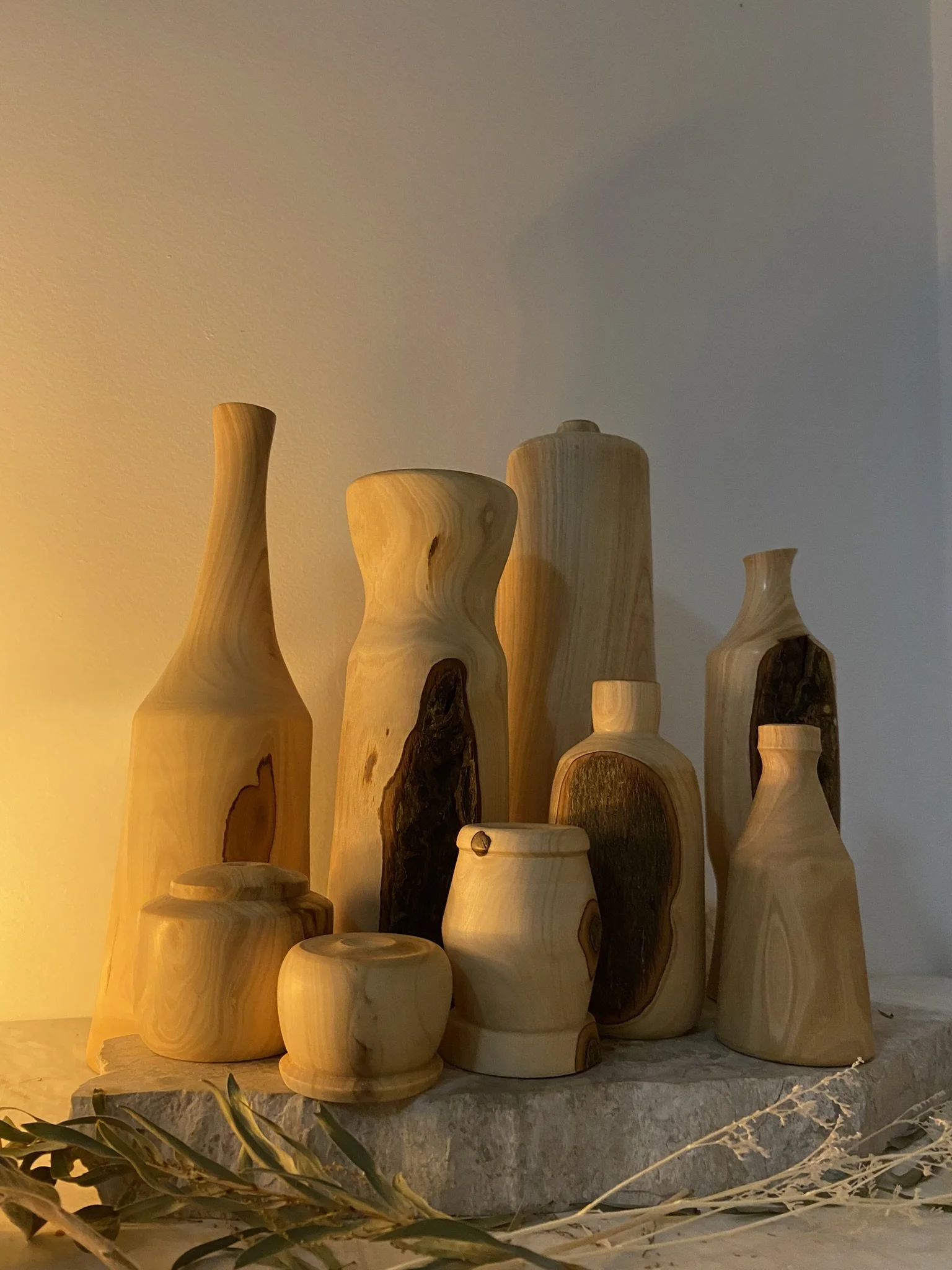 Collection of wooden vases and jars of various shapes and sizes on a stone surface with dried plant leaves in front.