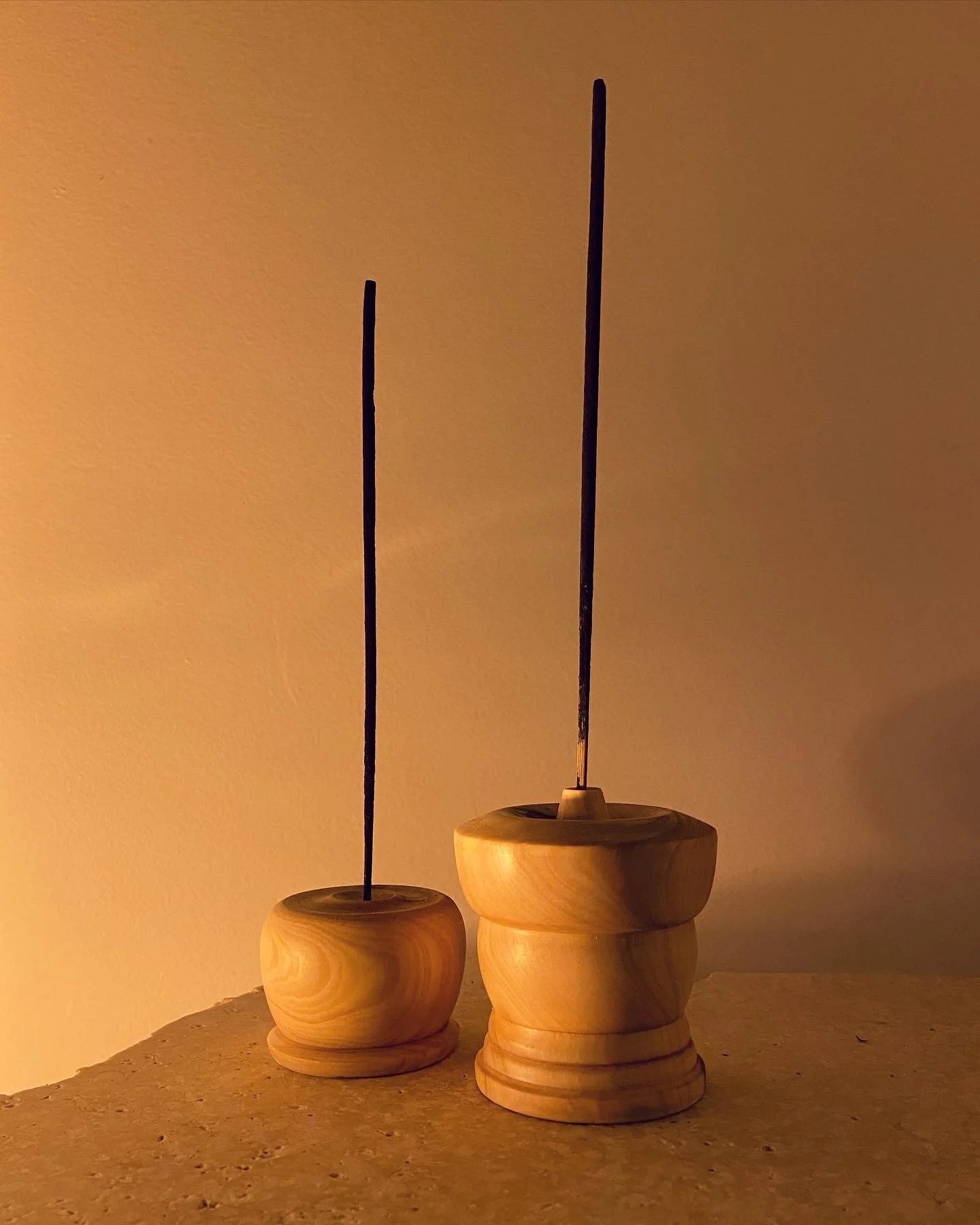 Two wooden incense holders with burning incense sticks on a textured surface against a plain wall