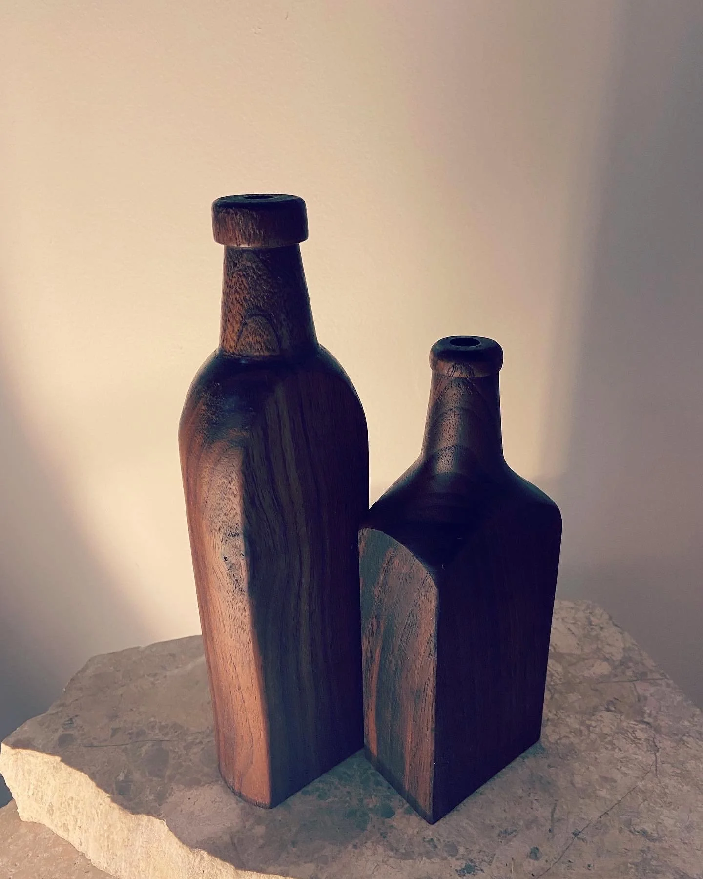 Two wooden bottles with narrow necks and round tops placed on a rough stone surface, against a plain light-colored wall.