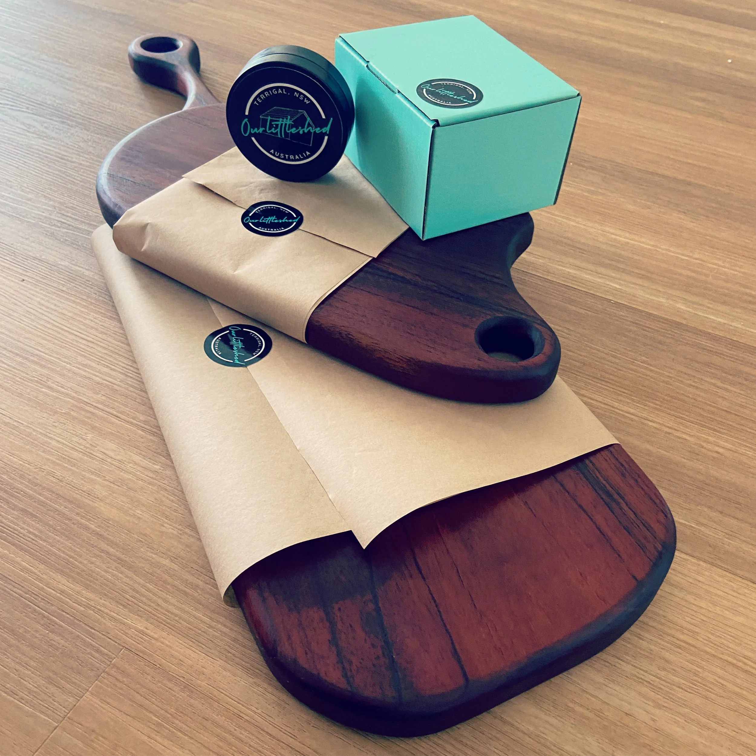 A wooden cutting board, a teal box, black container, and gift wrapped in brown paper with stickers on a wooden surface.