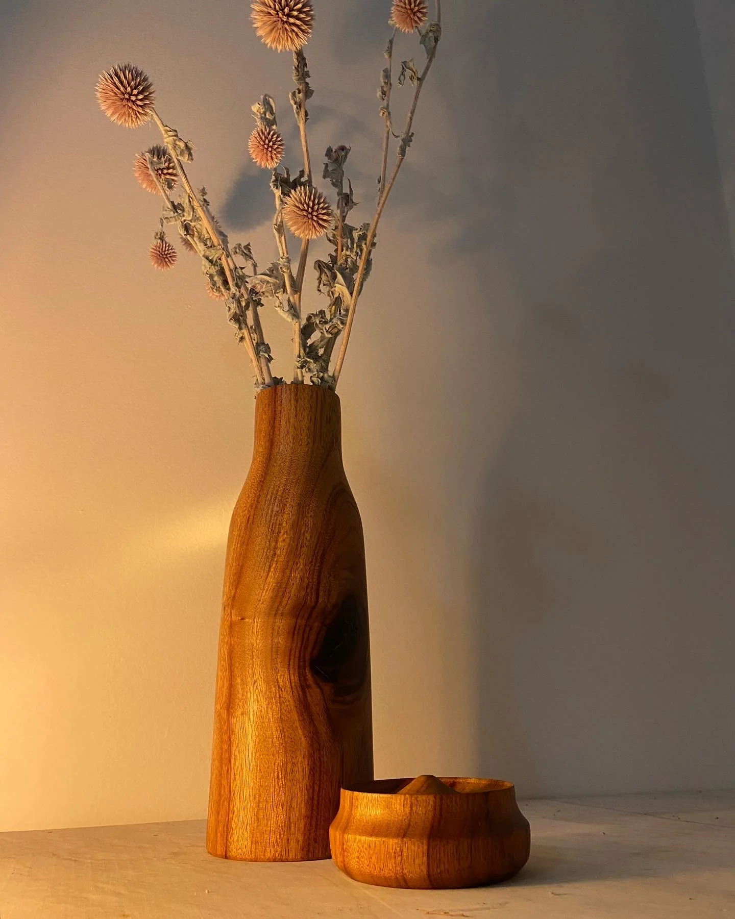 A tall wooden vase with dried flowers and a smaller wooden bowl on a light-colored surface against a plain wall.
