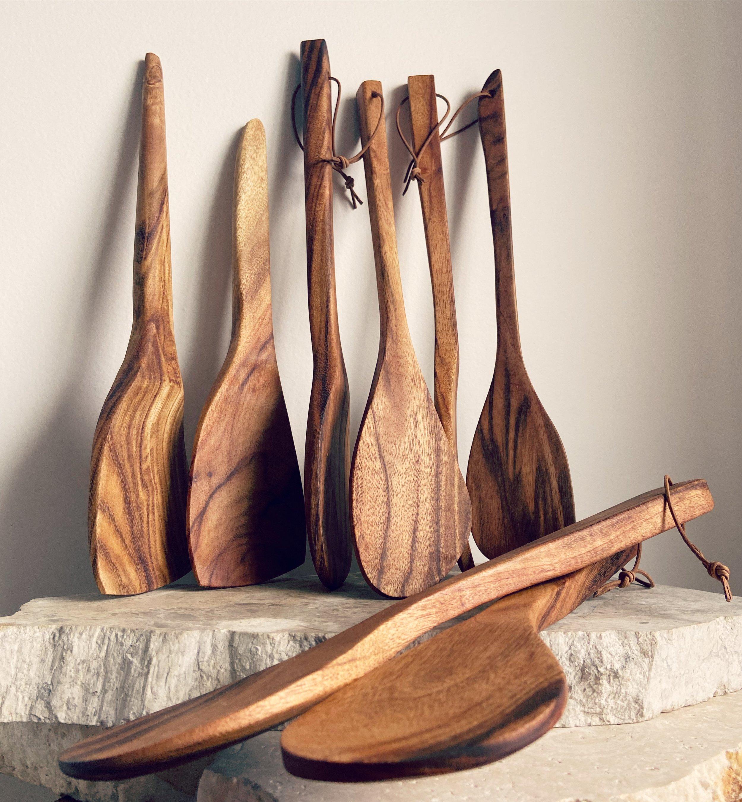 Set of six wooden spatulas with leather ties on a stone surface against a plain wall.