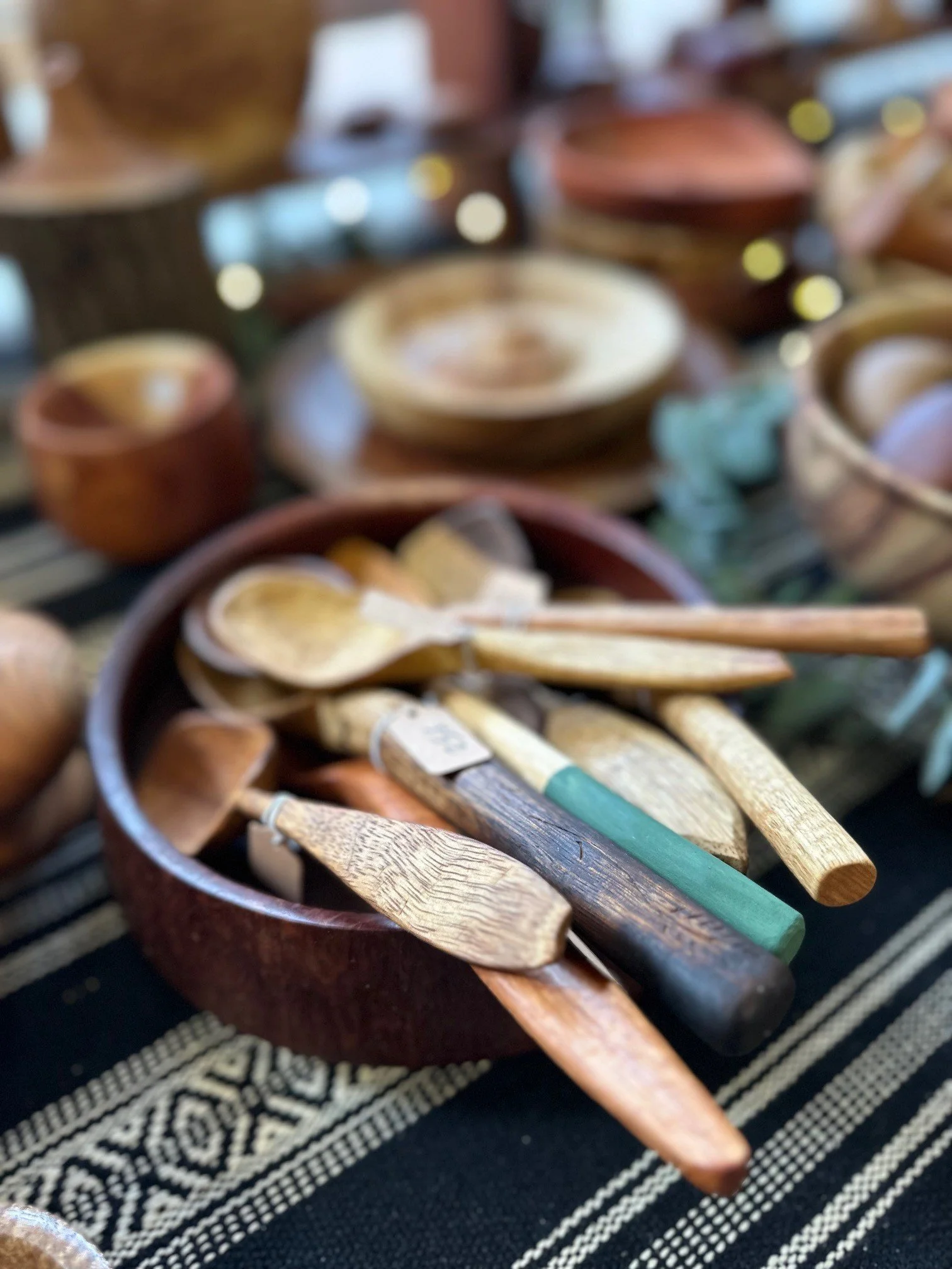Wooden spoons and utensils in a round bowl, with pottery items in the background, displayed on a striped cloth.