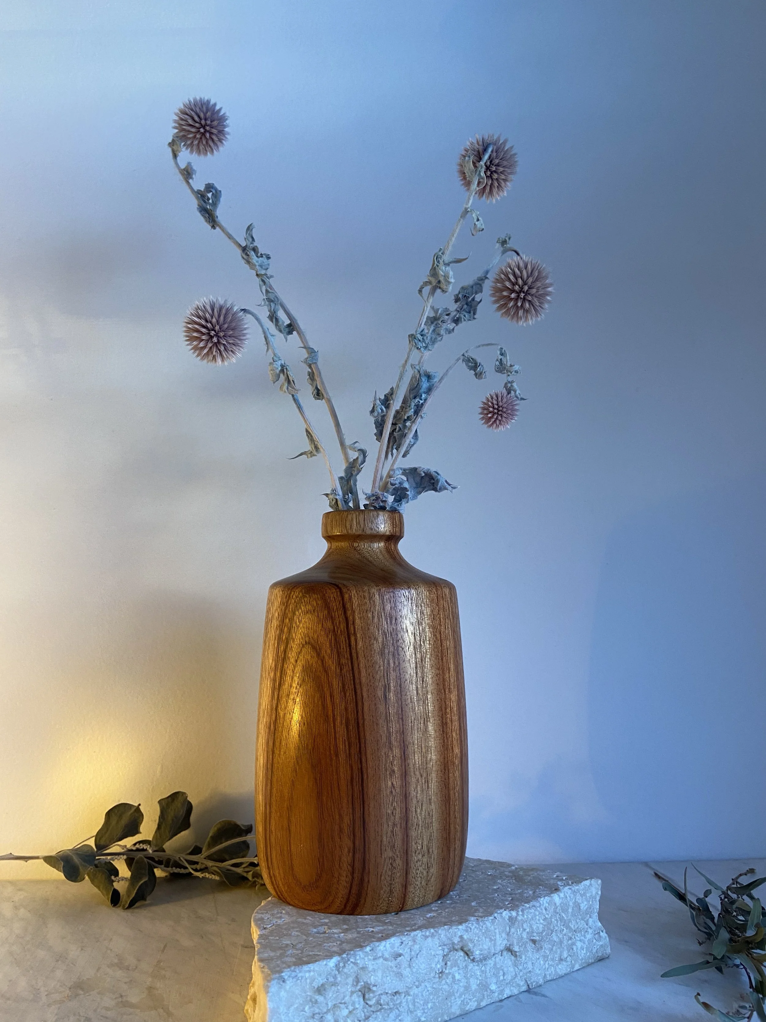 A wooden vase with dried flowers on a stone platform, with leaves and branches nearby, against a light and blue wall background.