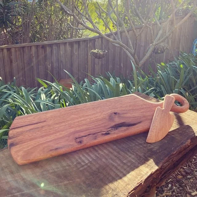 A wooden serving tray with a handle, placed on a wooden surface outdoors with greenery and a garden fence in the background.