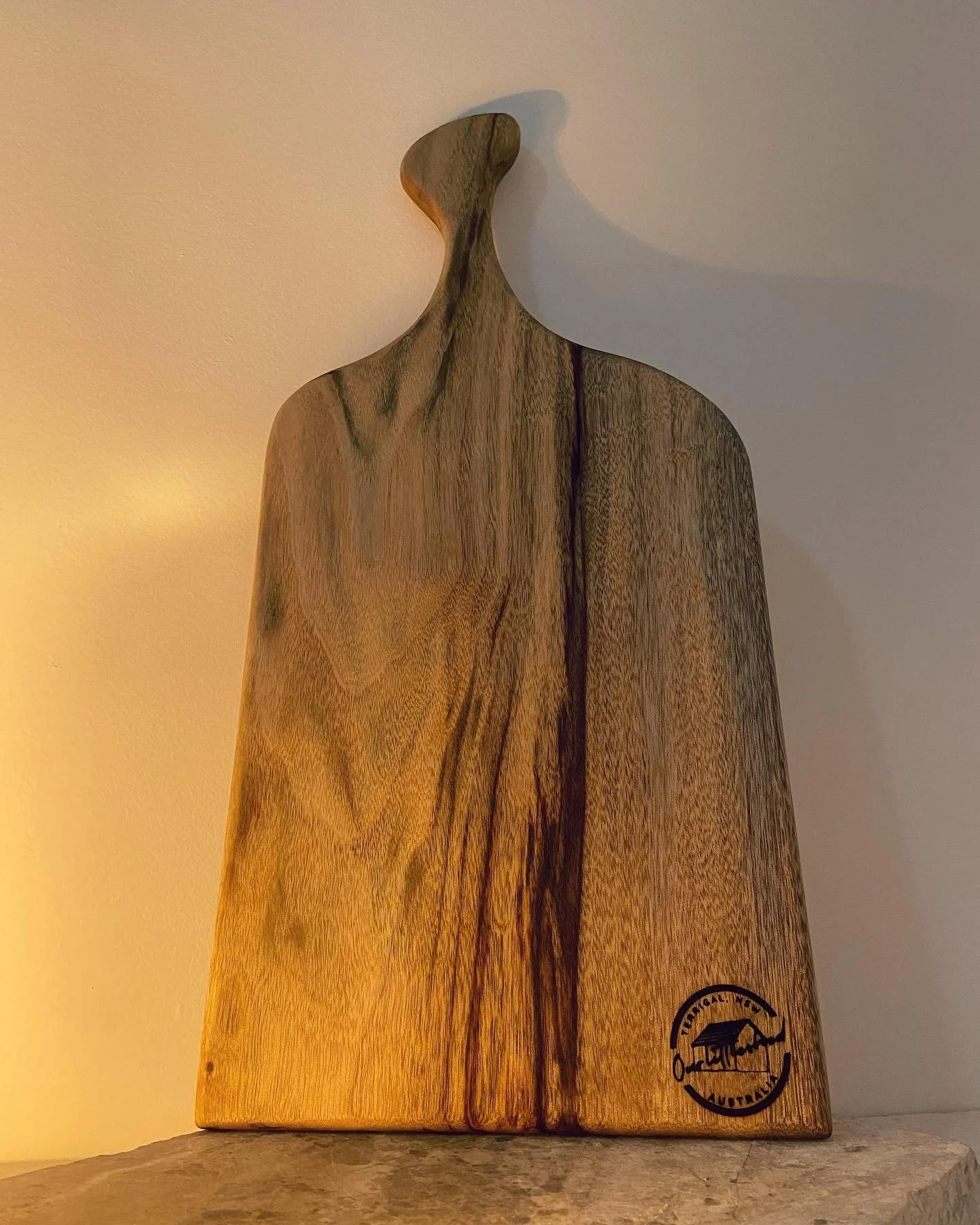 Wooden sculpture resembling a person with an elongated head, placed on a stone surface, with a circular logo near the bottom right corner.
